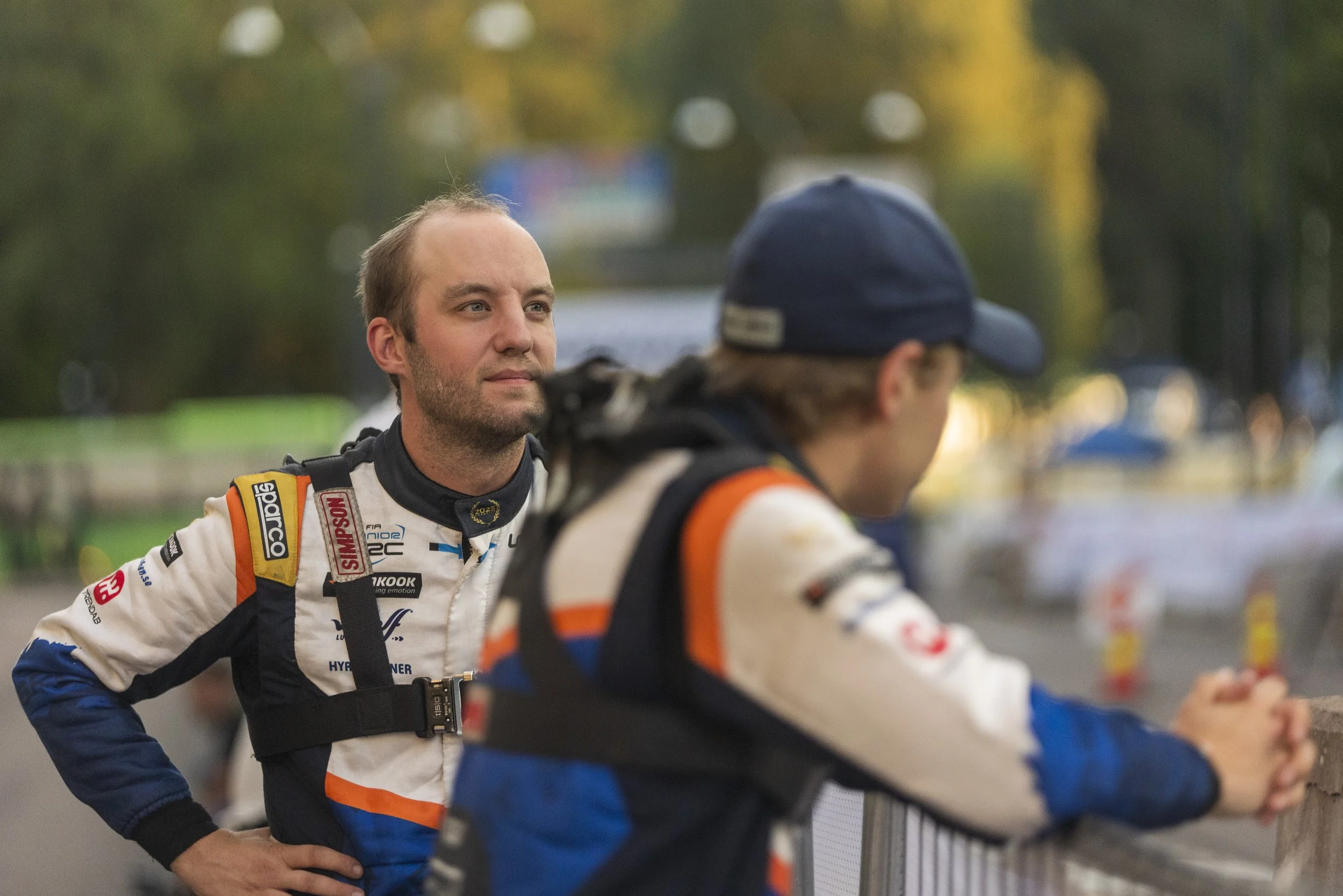 A race car driver in a racing suit standing and talking to another person in a racing suit in a paddock area with trees in the background.