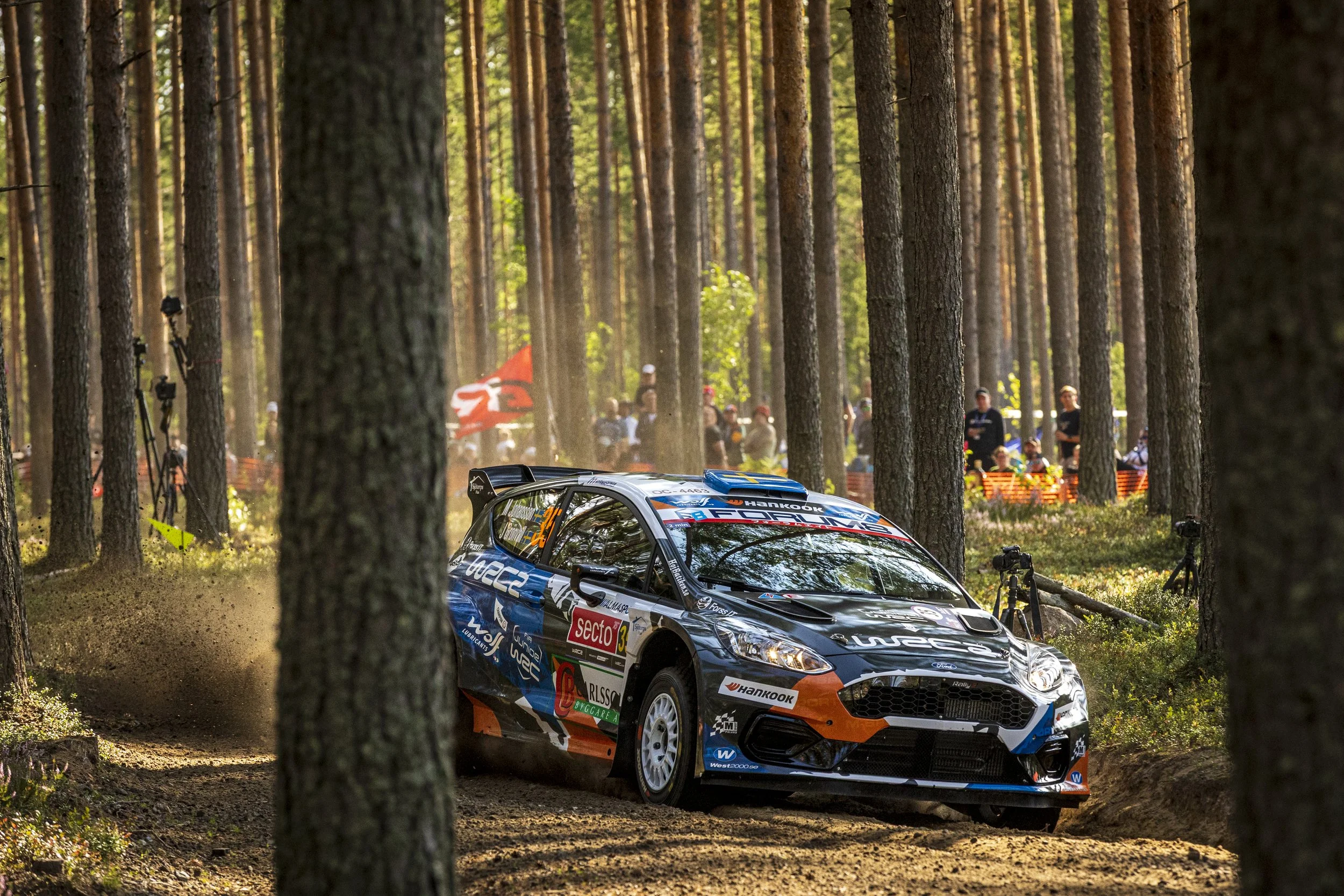 A rally car racing through a forested dirt trail, kicking up dust, with spectators and cameras in the background.