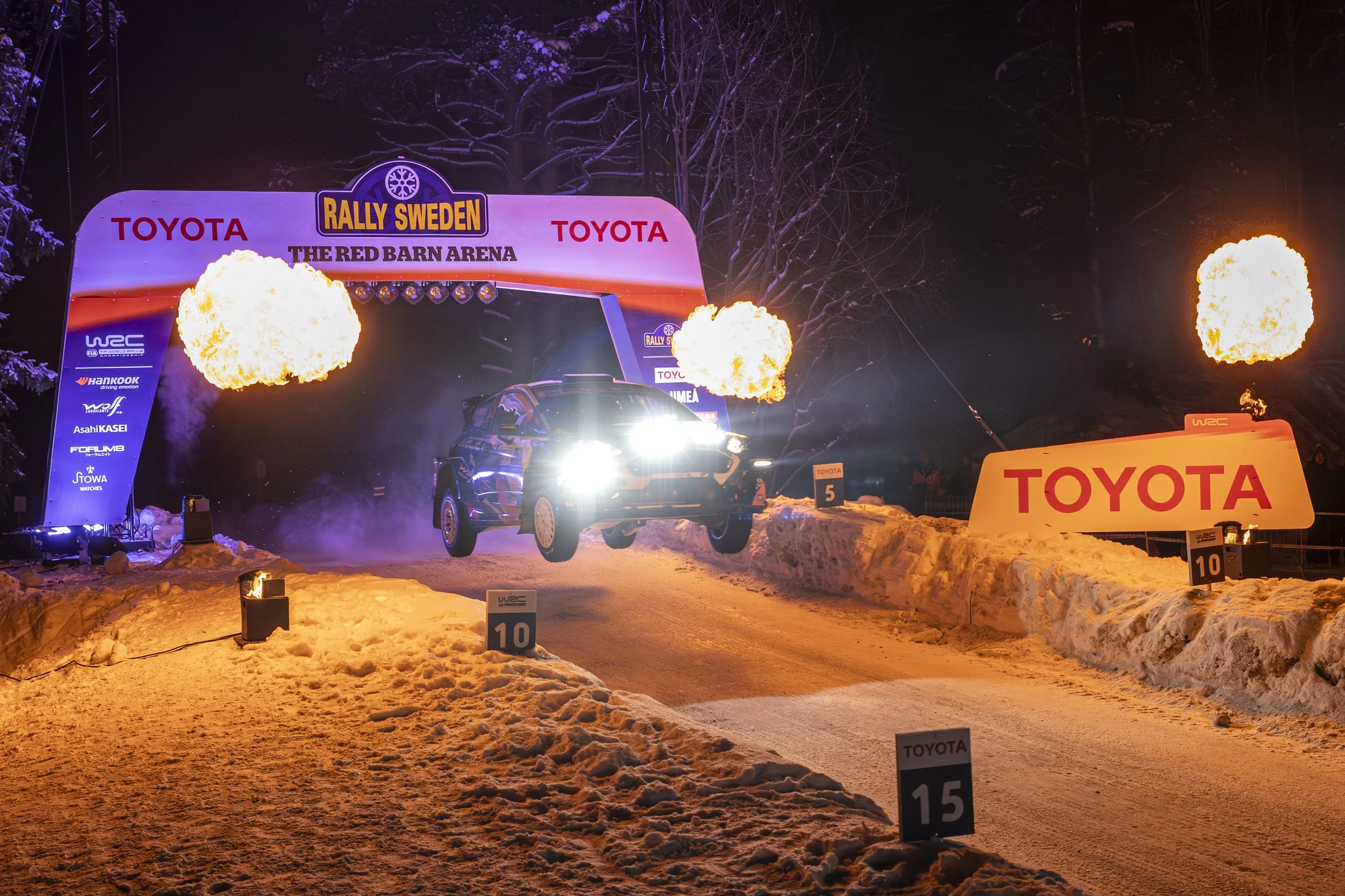 Nighttime rally car race in snowy terrain with flames, illuminated signs, and a rally car crossing the finish line.