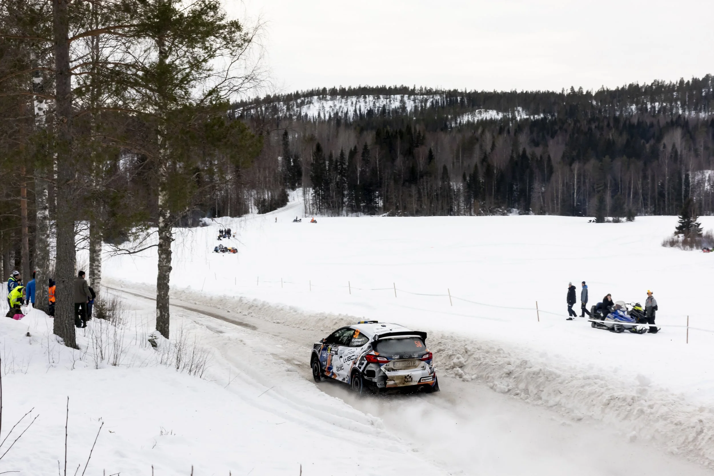 Snow-covered landscape with a rally car driving on a snowy road, spectators standing near the road watching, and snow mobile riders in the background. Tall trees and a forested hillside are visible in the distance.