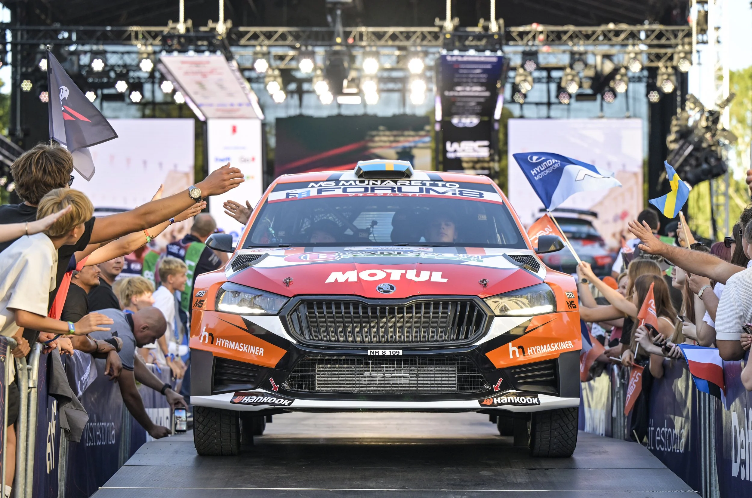 A rally car on a stage with spectators reaching out to it, flags waving, and bright lights and screens in the background.