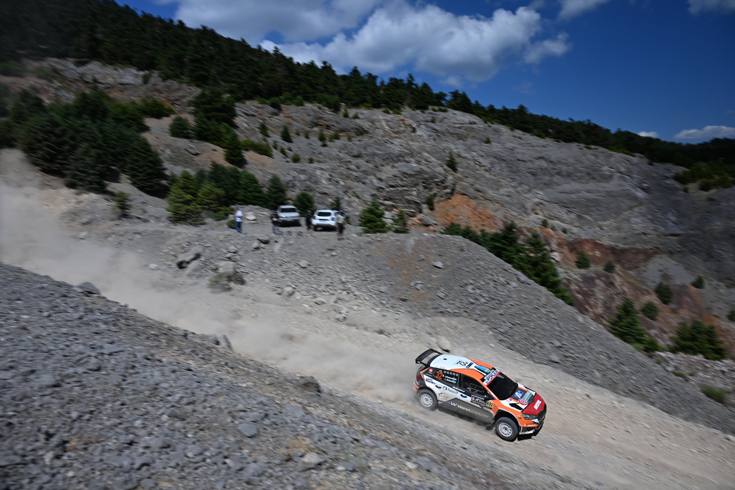 A rally car racing on a dirt mountain road, with rocky slopes and pine trees in the background under a blue sky with clouds.