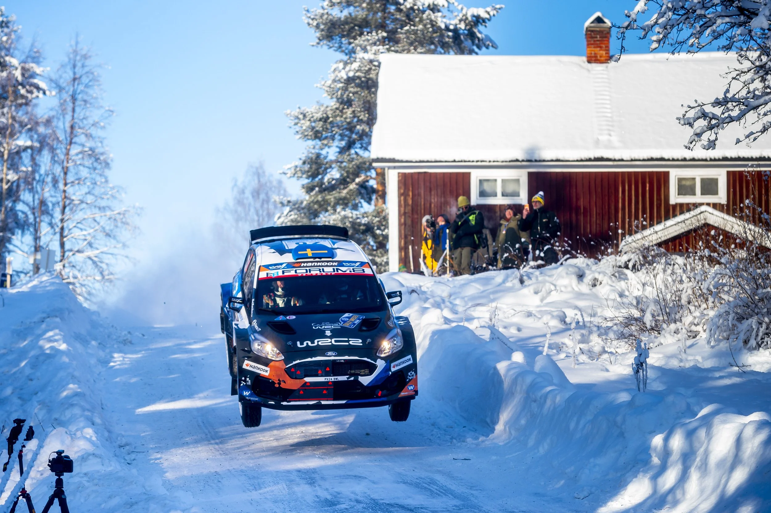 A rally car racing on a snow-covered road with spectators and a house in the background, surrounded by winter trees.