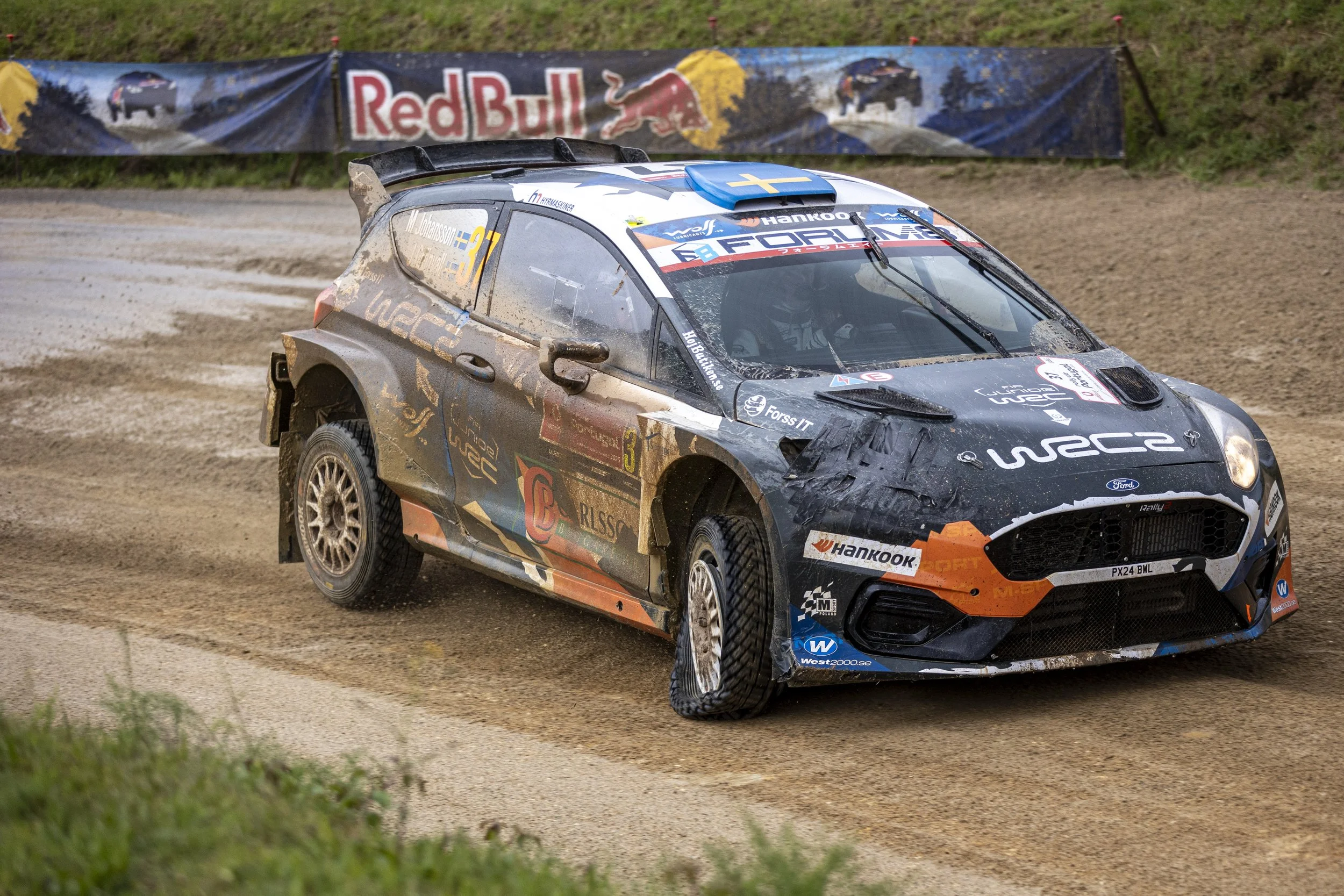 A rally car on a dirt track during a race, with a Red Bull banner in the background.