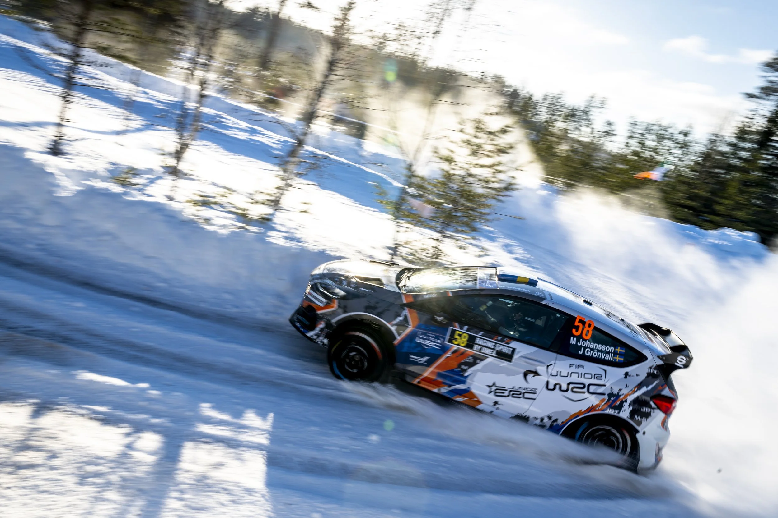 A rally car in motion on a snowy forest track, with trees and snow visible in the background, and dust or snow spray behind it.