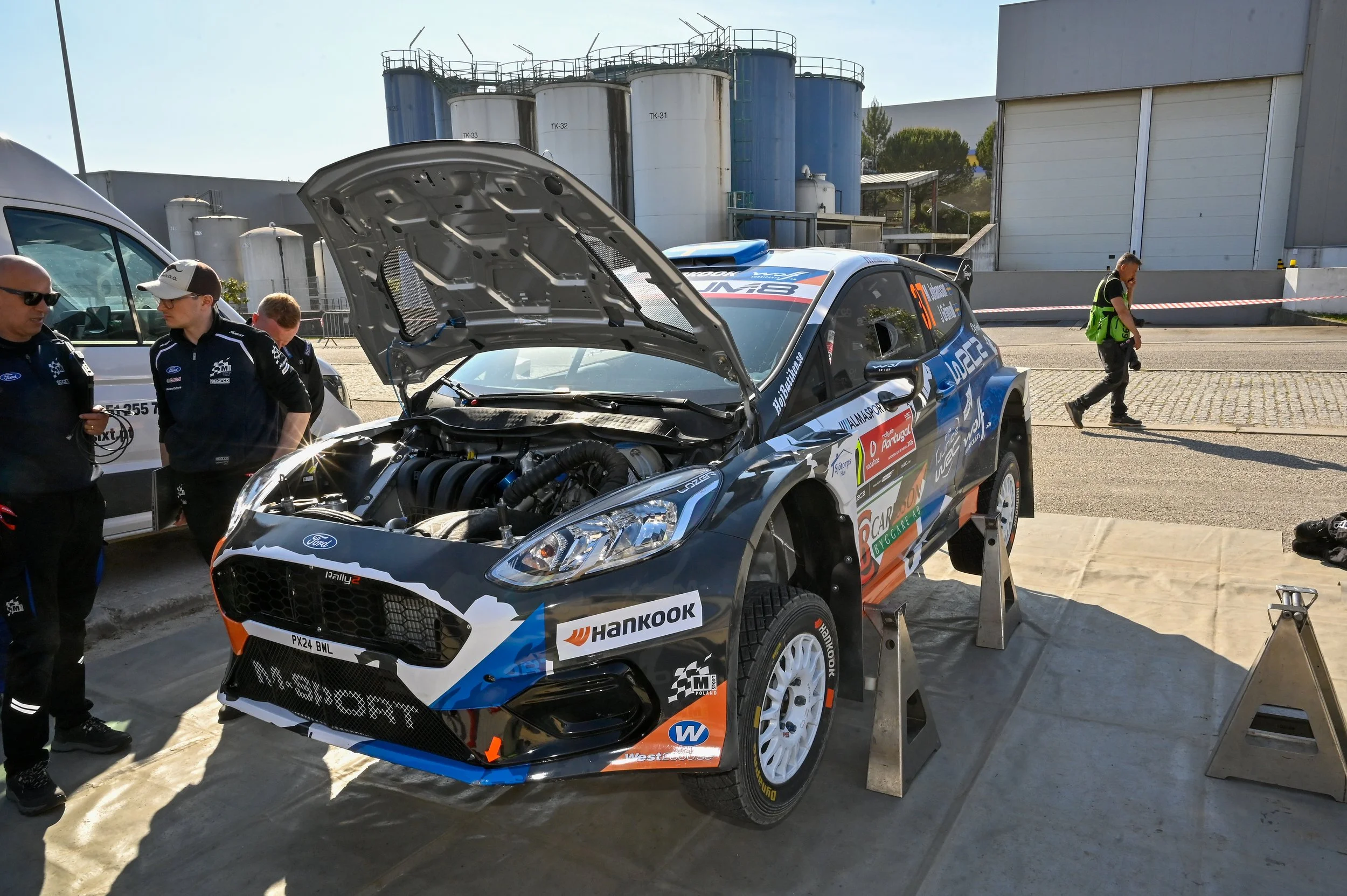 A rally car with its hood open is being inspected or repaired, with team members and a person walking nearby in an industrial area or service zone.