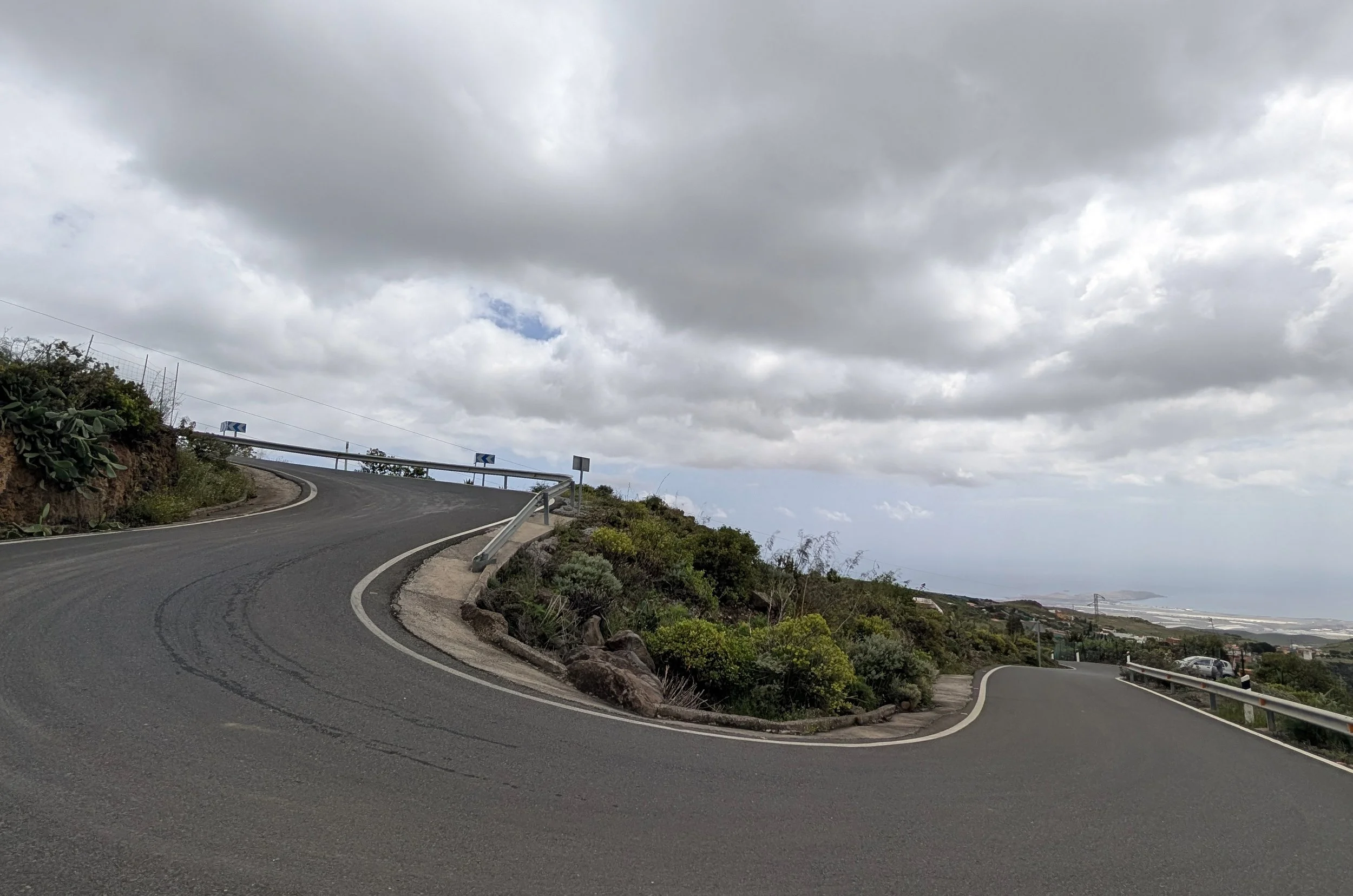 A winding mountain road with guardrails on the side, rocky terrain with sparse vegetation, and a cloudy sky overhead.