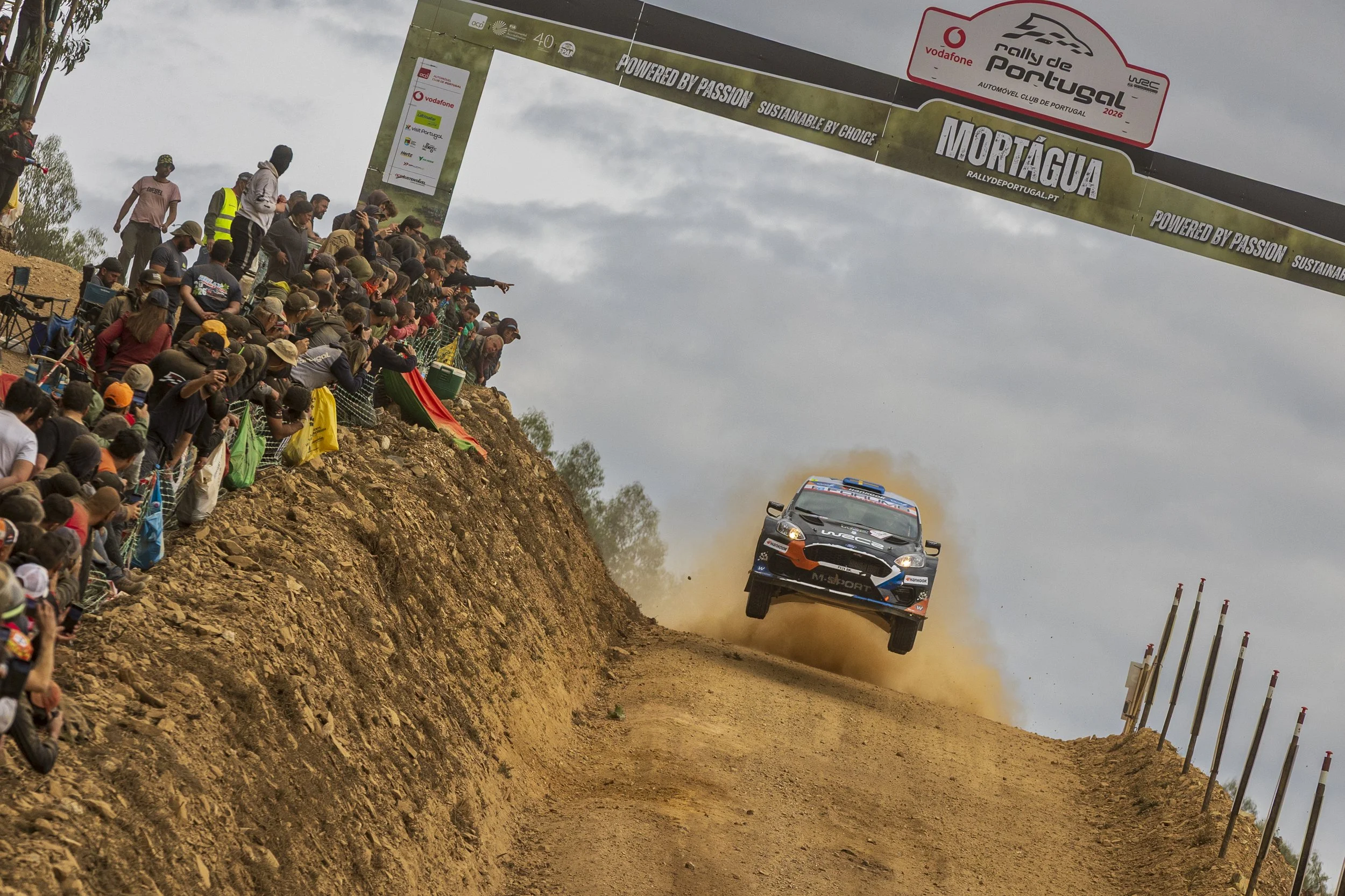 A rally car flying through the air on a dirt track with a crowd of spectators watching from the hillside and a large banner overhead.