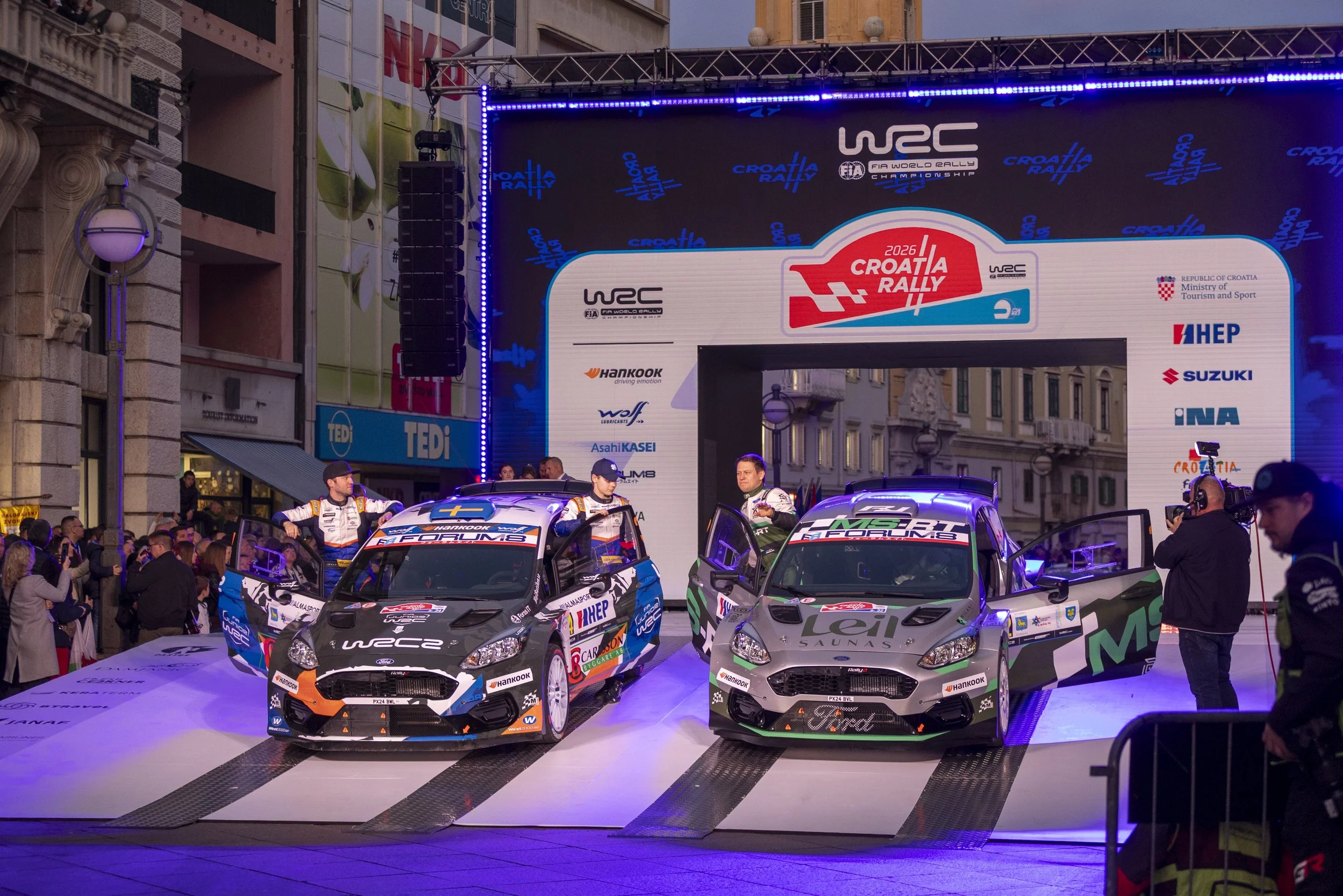 Three rally cars on display at the 2026 Croatia Rally, with people and spectators surrounding them. The backdrop features a large digital screen with logos of WRC, Croatia Rally, and various sponsors.