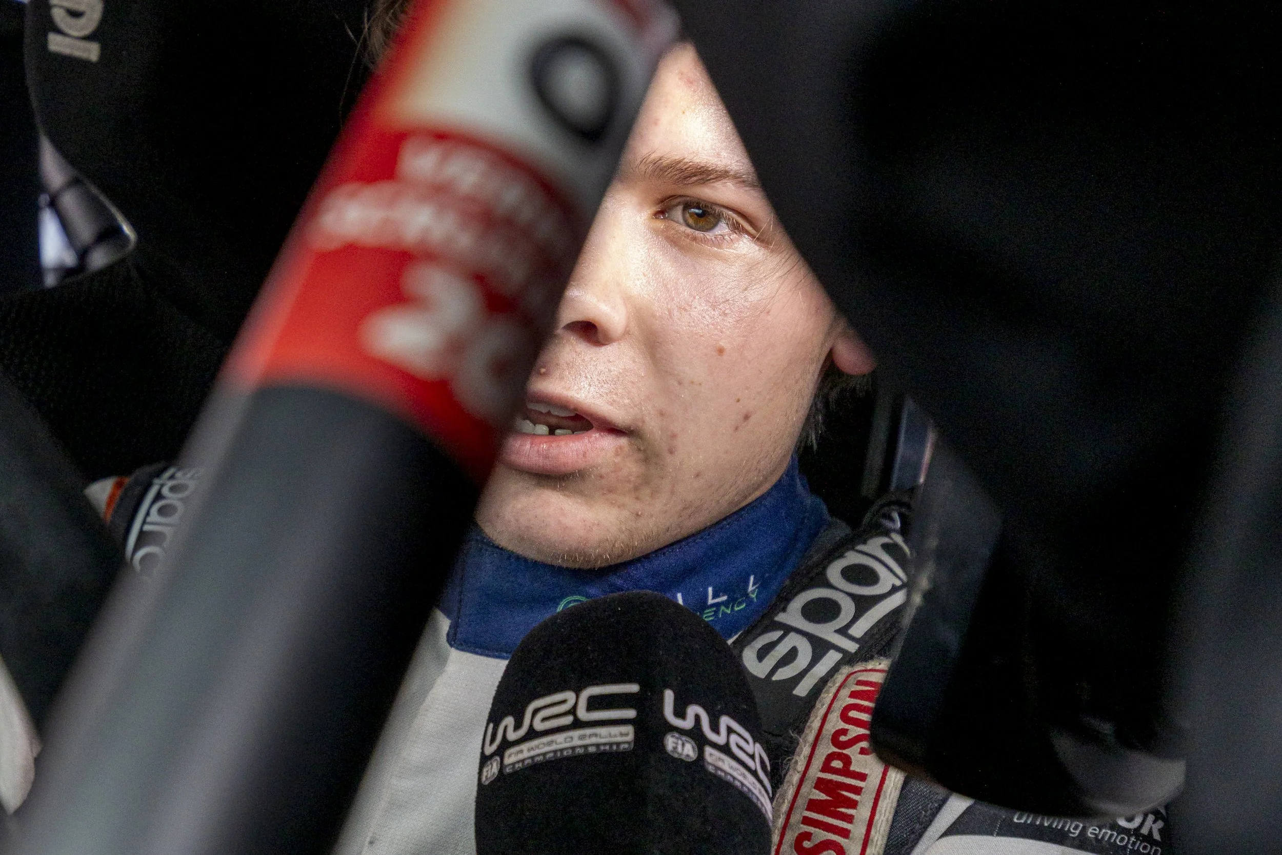 A race car driver with face paint and racing gear, sitting in a vehicle, partially obscured by equipment, looking ahead.