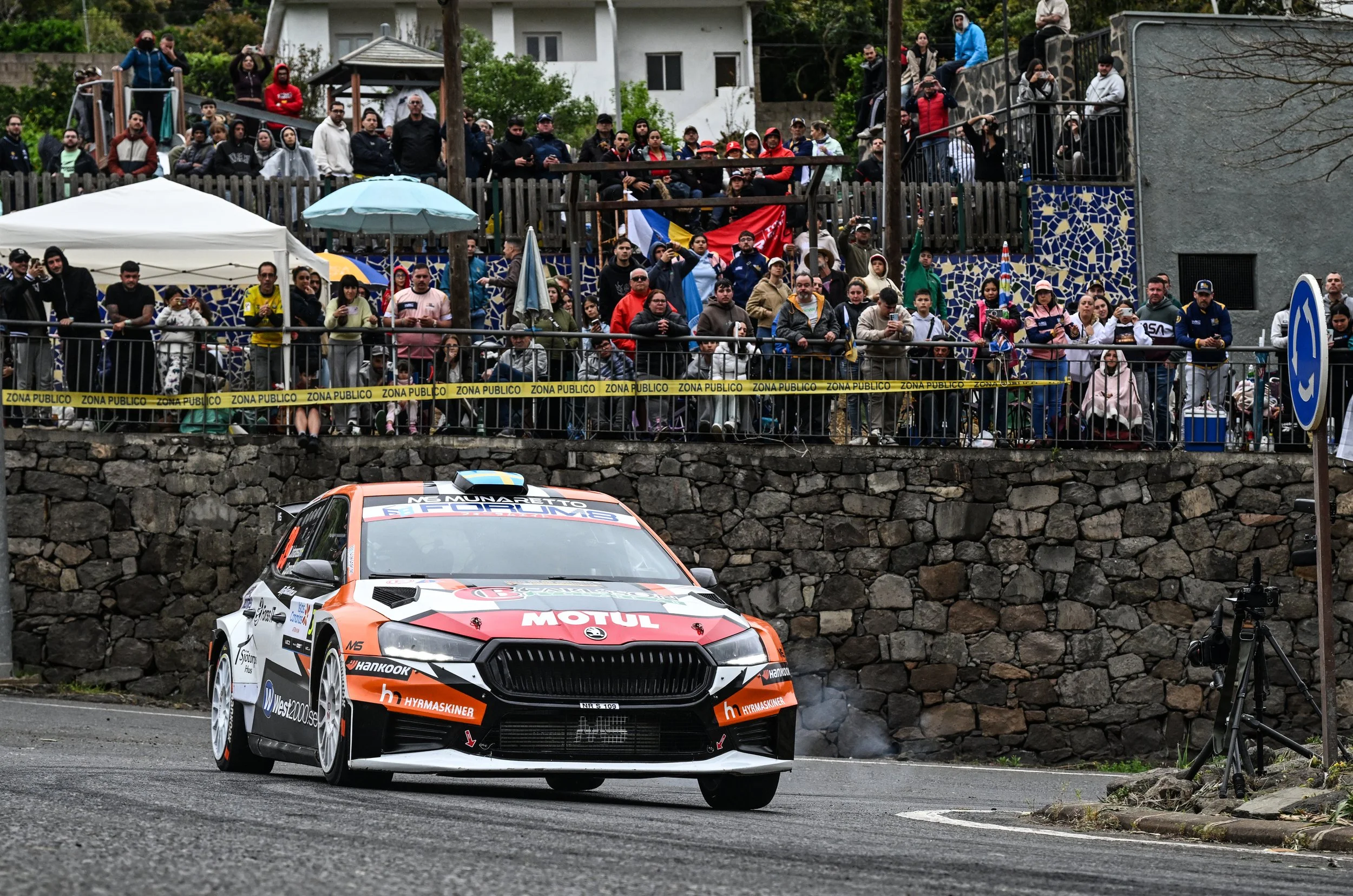 A rally car on a road with a crowd of spectators behind a safety barrier, some holding umbrellas, watching the race.