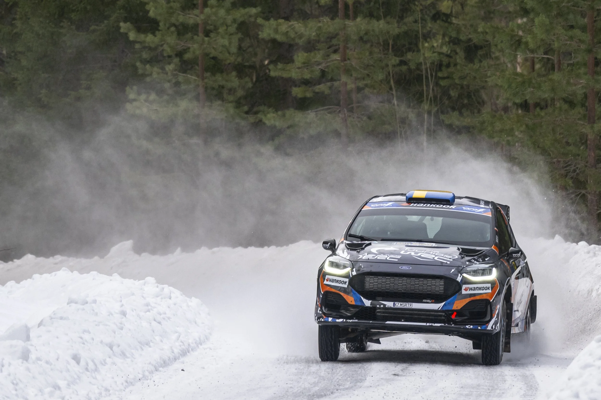 A black rally car driving on a snow-covered road through a snowy forest with tall evergreen trees in the background, kicking up snow and mist.