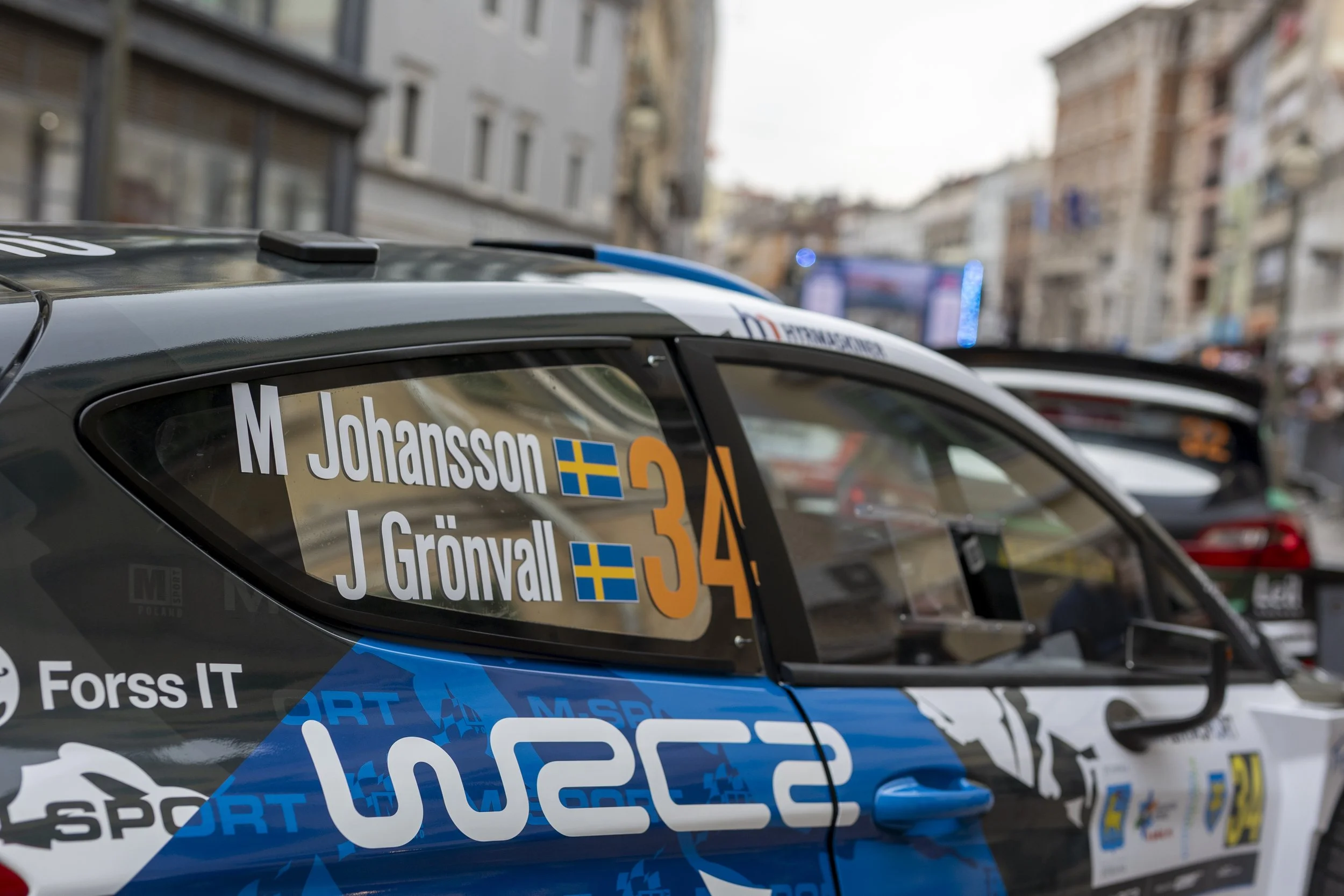 Race cars lined up on a city street, with helmets and racing decals, indicating a rally or racing event.