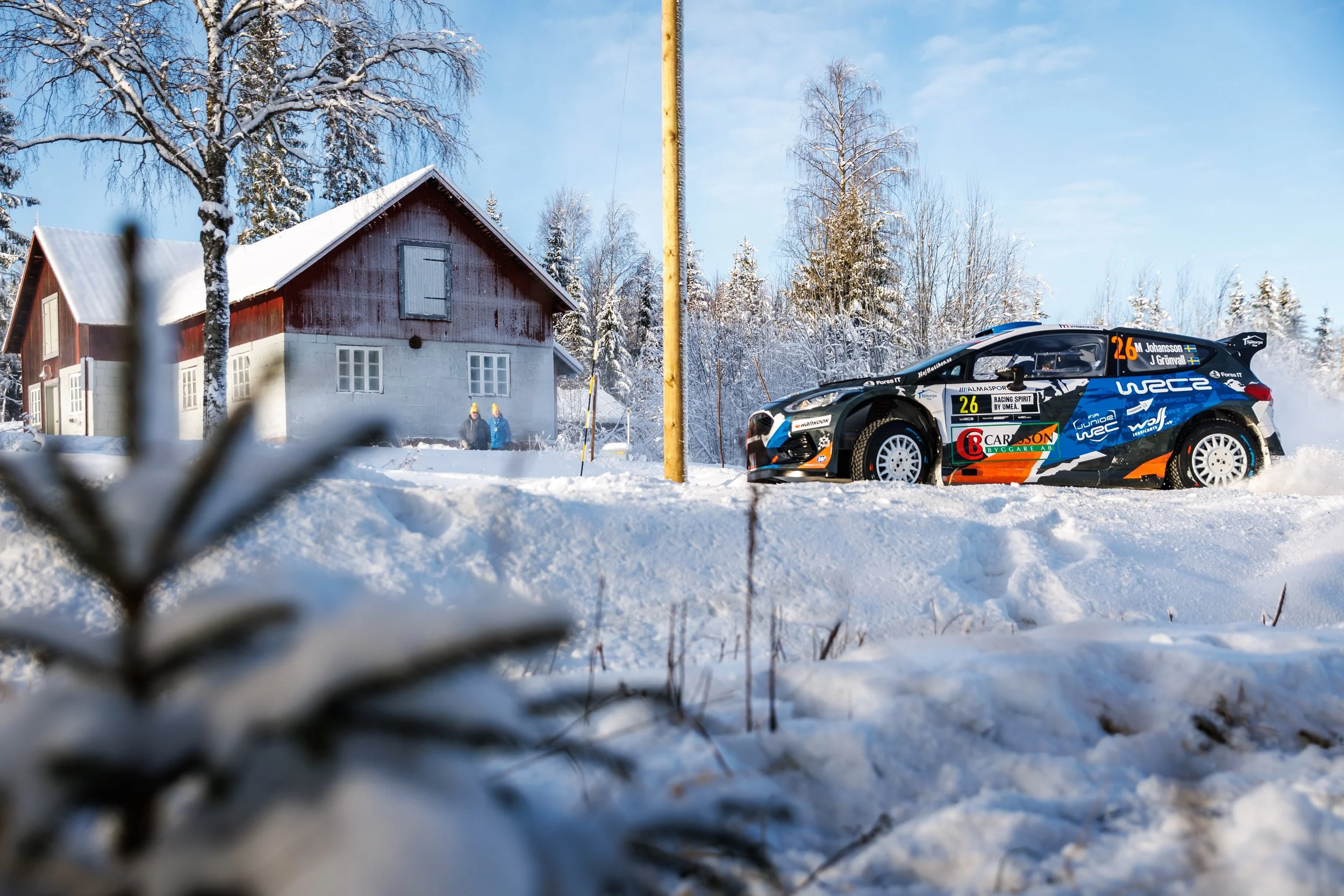 Snow-covered landscape with a house, trees, a utility pole, and a rally car with the number 26, Swedish flags, and racing sponsor logos.