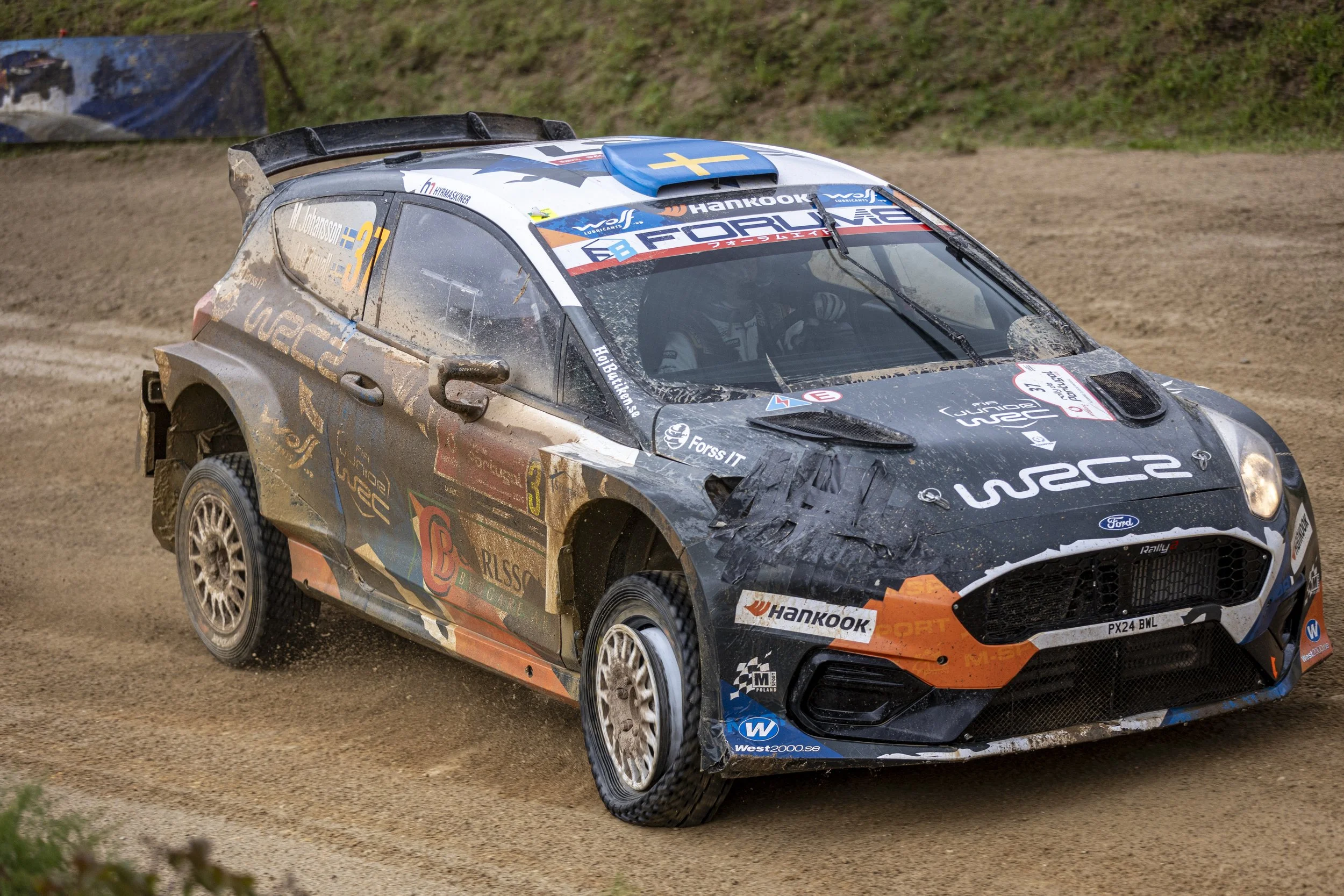 A rally car driving on a dirt track, kicking up dust, with the number 37 on the side, decorated with sponsor logos and a Swedish flag on the roof.