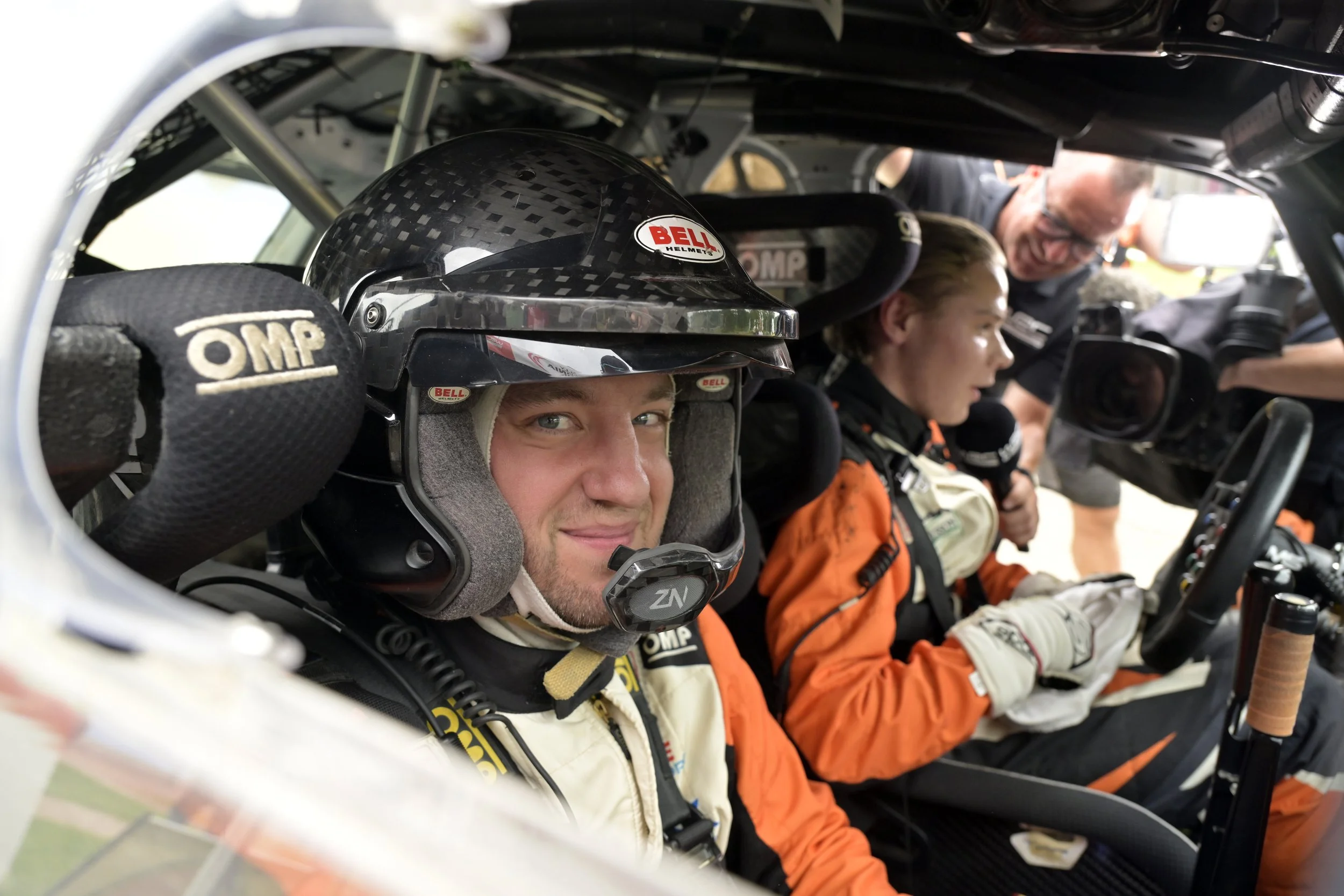Race car drivers inside the cockpit, with one driver in focus wearing a racing helmet and suit, with team members attending to the driver and the car's controls.