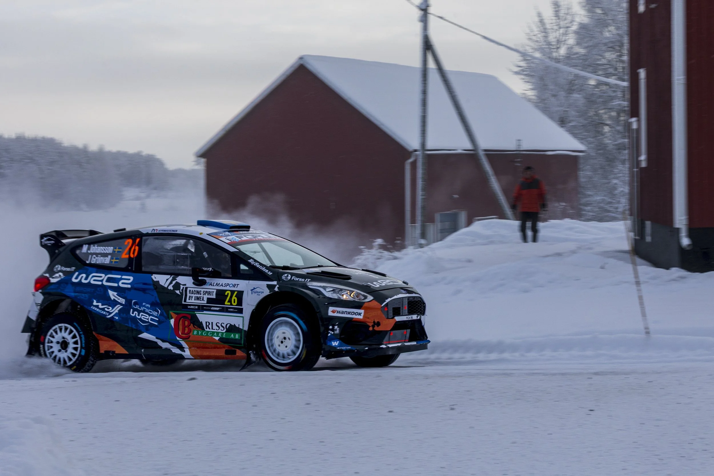 A rally car racing on a snow-covered road, with snow and a red building in the background, and a person standing nearby.