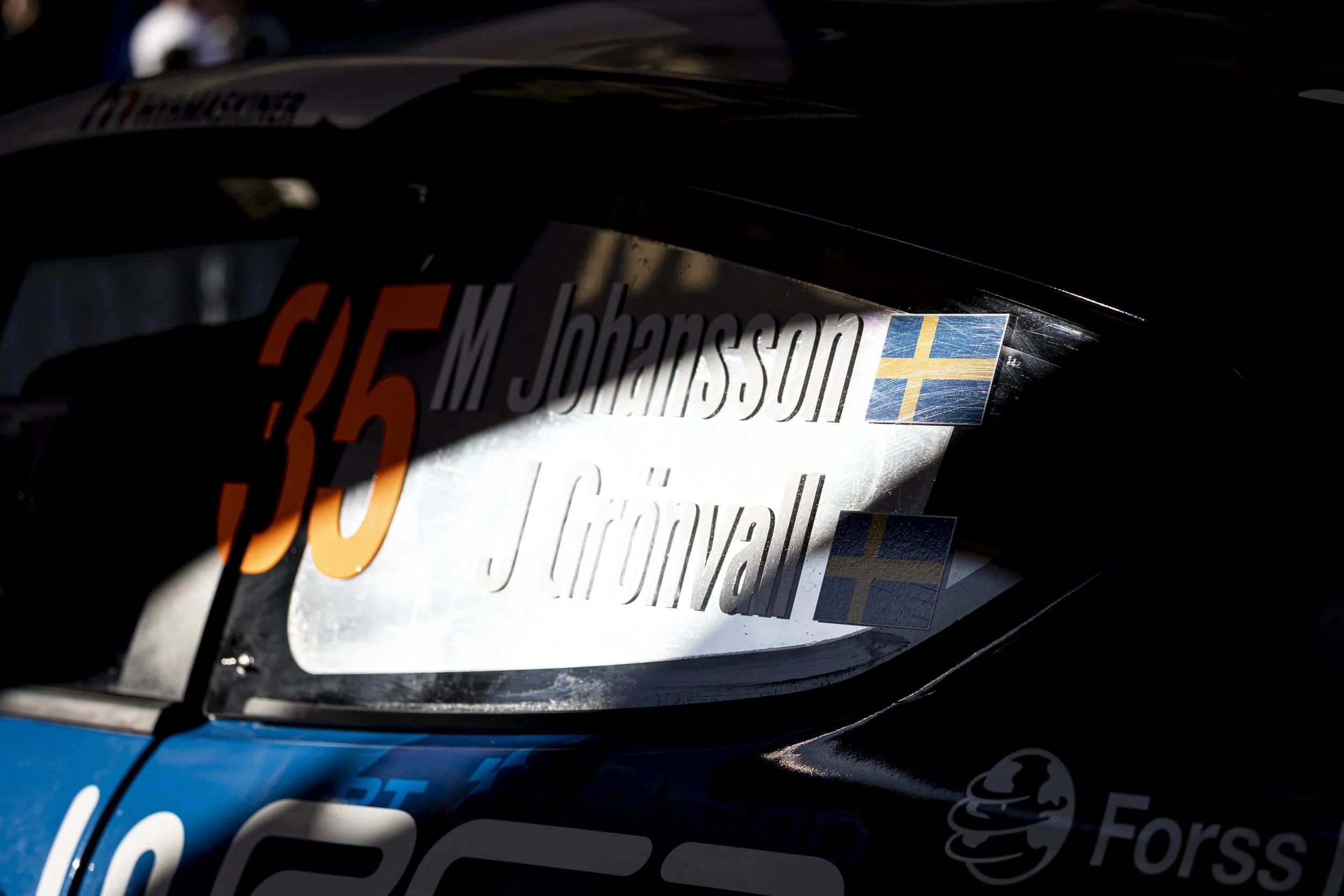 Close-up of a racing car's hood with decals, including the name 'M. Johansson' and the Swedish flag, partially illuminated.