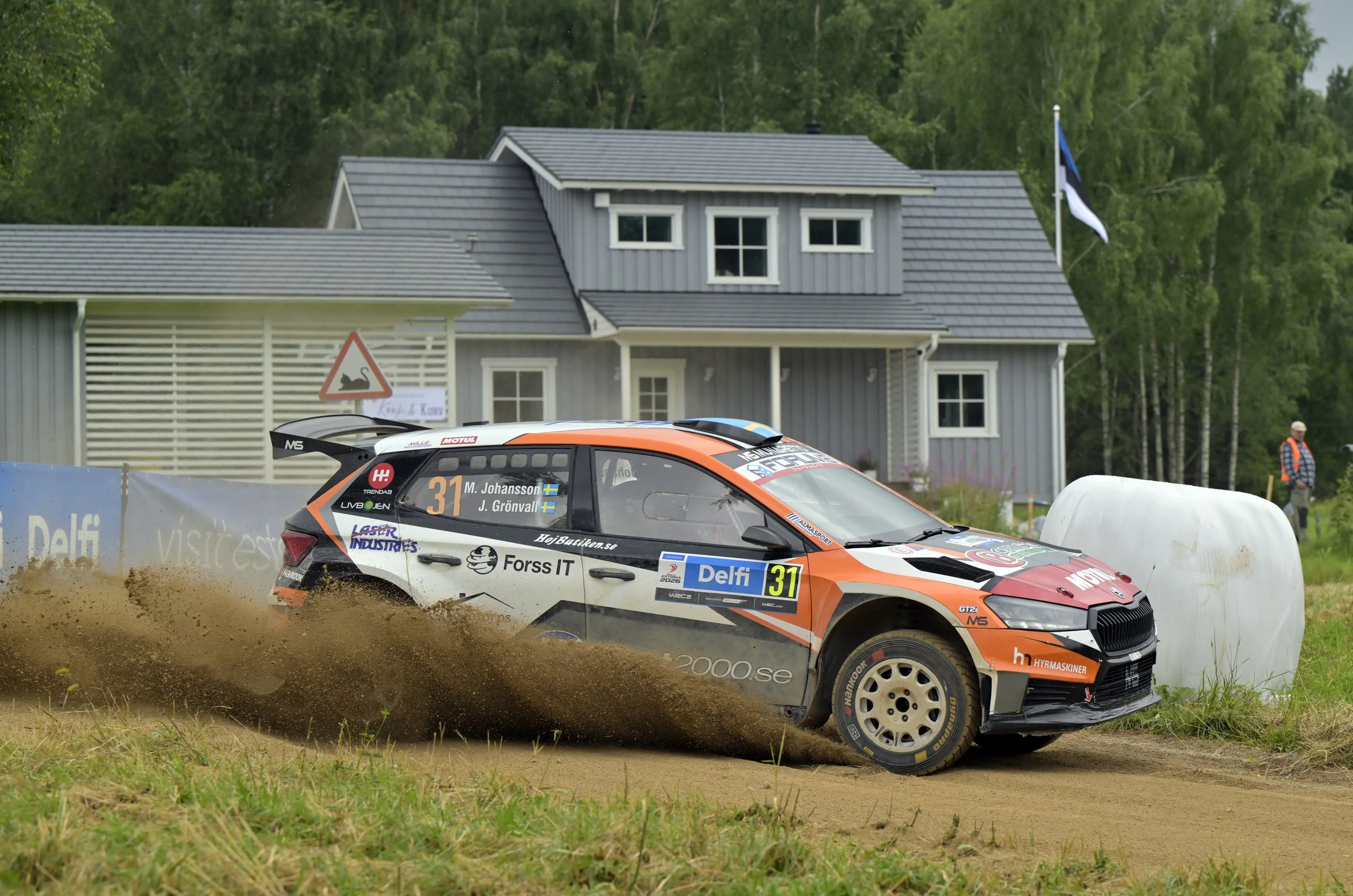 A rally car is racing on a dirt track, kicking up dirt as it goes. The car is black and orange with various sponsor logos, and has the number 31 on it. In the background, there is a gray house with white trim, surrounded by green trees and a blue fla