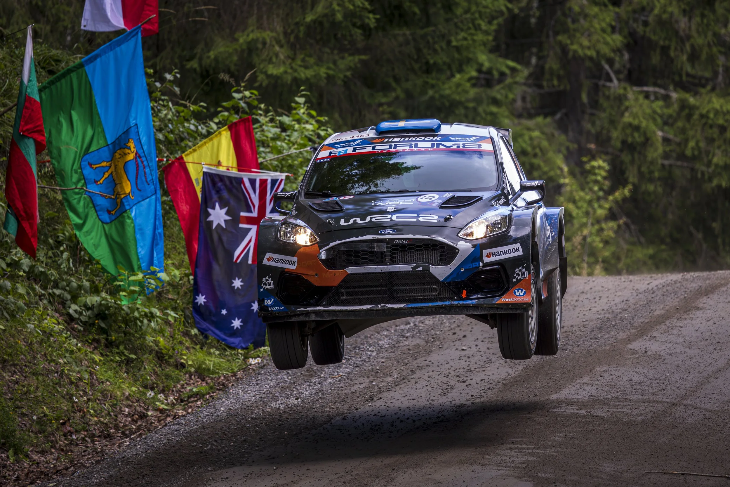 A black rally car on a dirt road surrounded by lush green trees, with colorful flags along the side of the road.