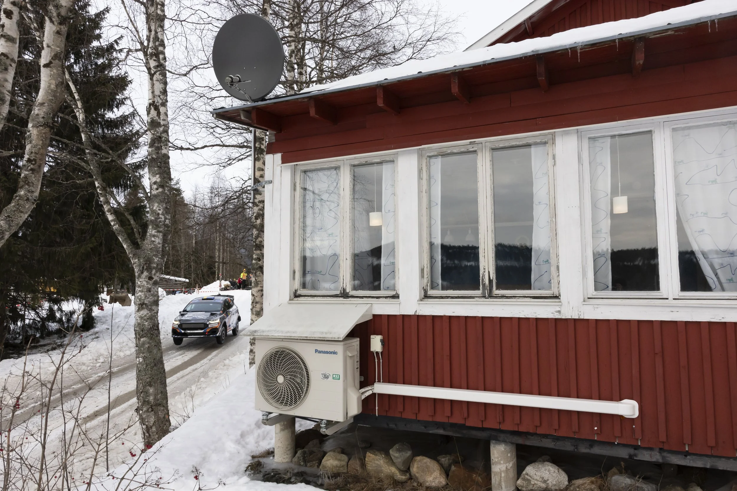 A red house with large windows, an air conditioning unit outside, and a satellite dish on the roof. There is snow on the ground and trees surrounding the house. A car is parked nearby.