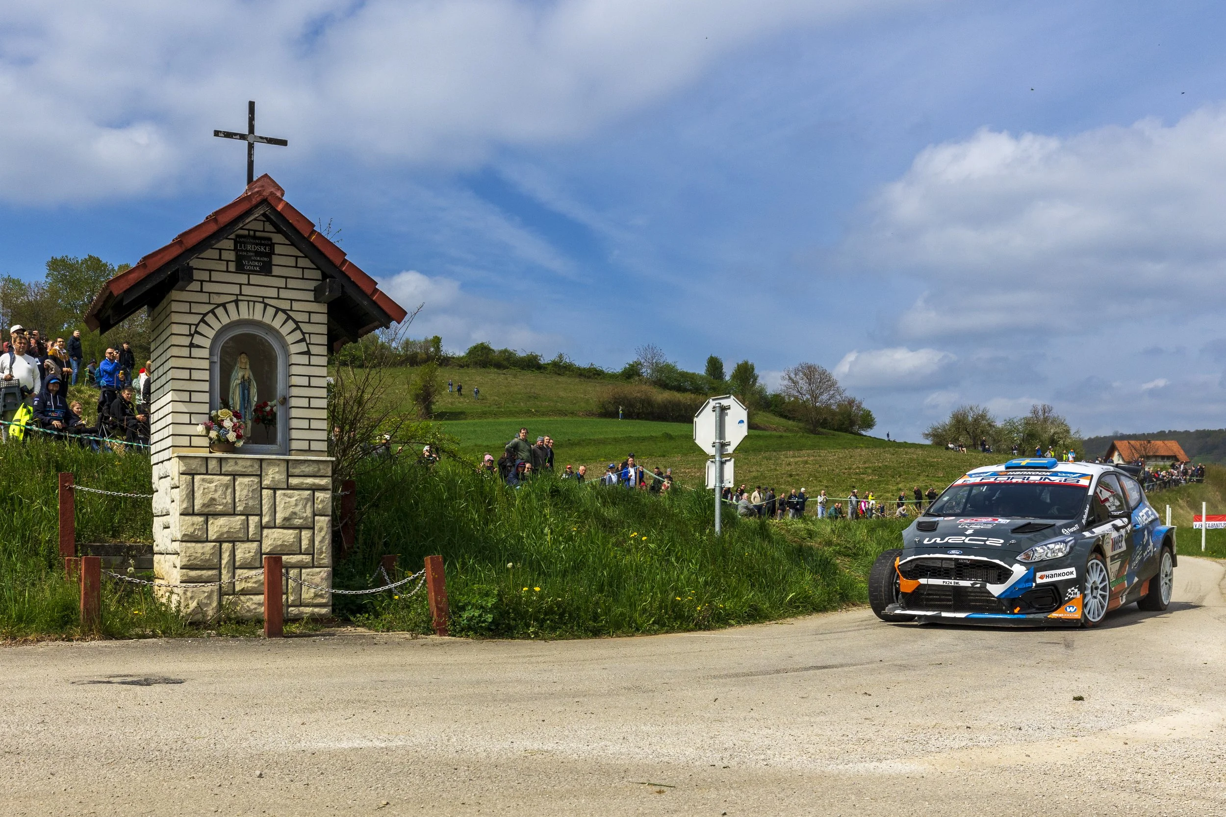 A small religious shrine with a statue of the Virgin Mary inside, decorated with flowers, on a grassy area near a dirt road. A crowd of people is gathered on a hillside in the background, and a rally car with sponsor logos is driving past in the fore