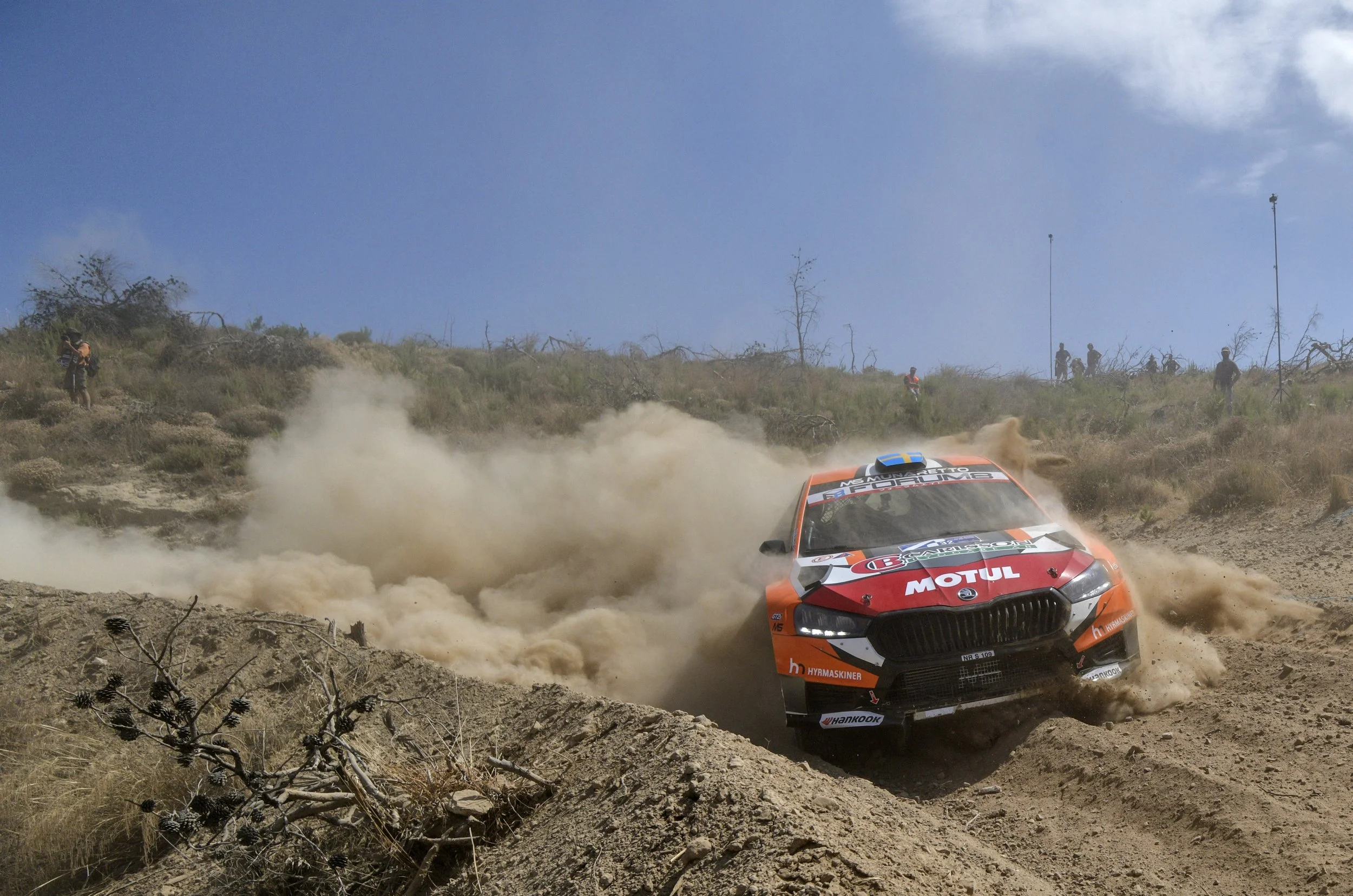 An orange rally car kicking up dust on a dirt rally track with a dry, shrubby landscape and a few spectators in the background.