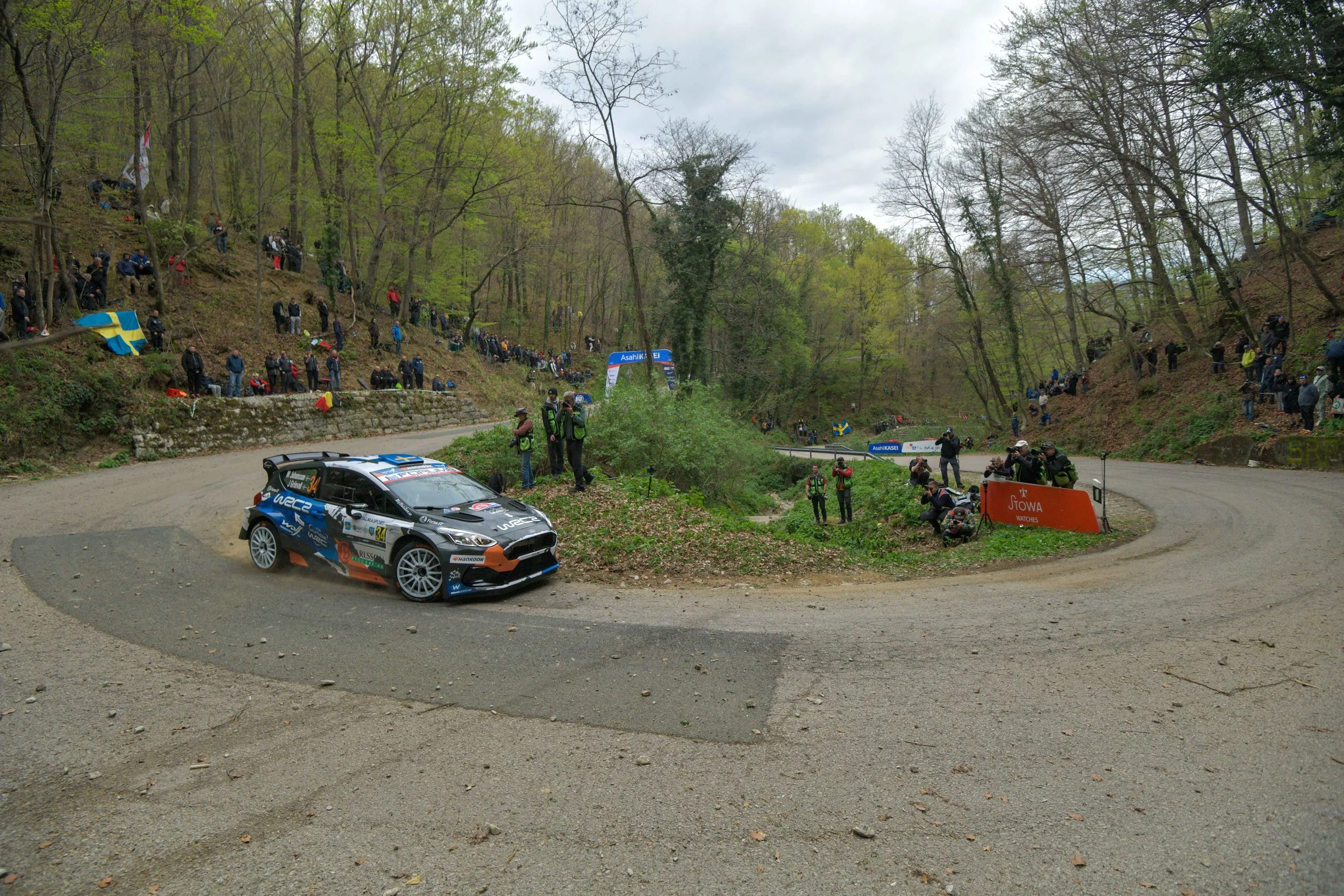 A rally car on a dirt road during a race, with spectators standing along the hillside and at the edge of the road, some holding flags, in a wooded area with cloudy sky.