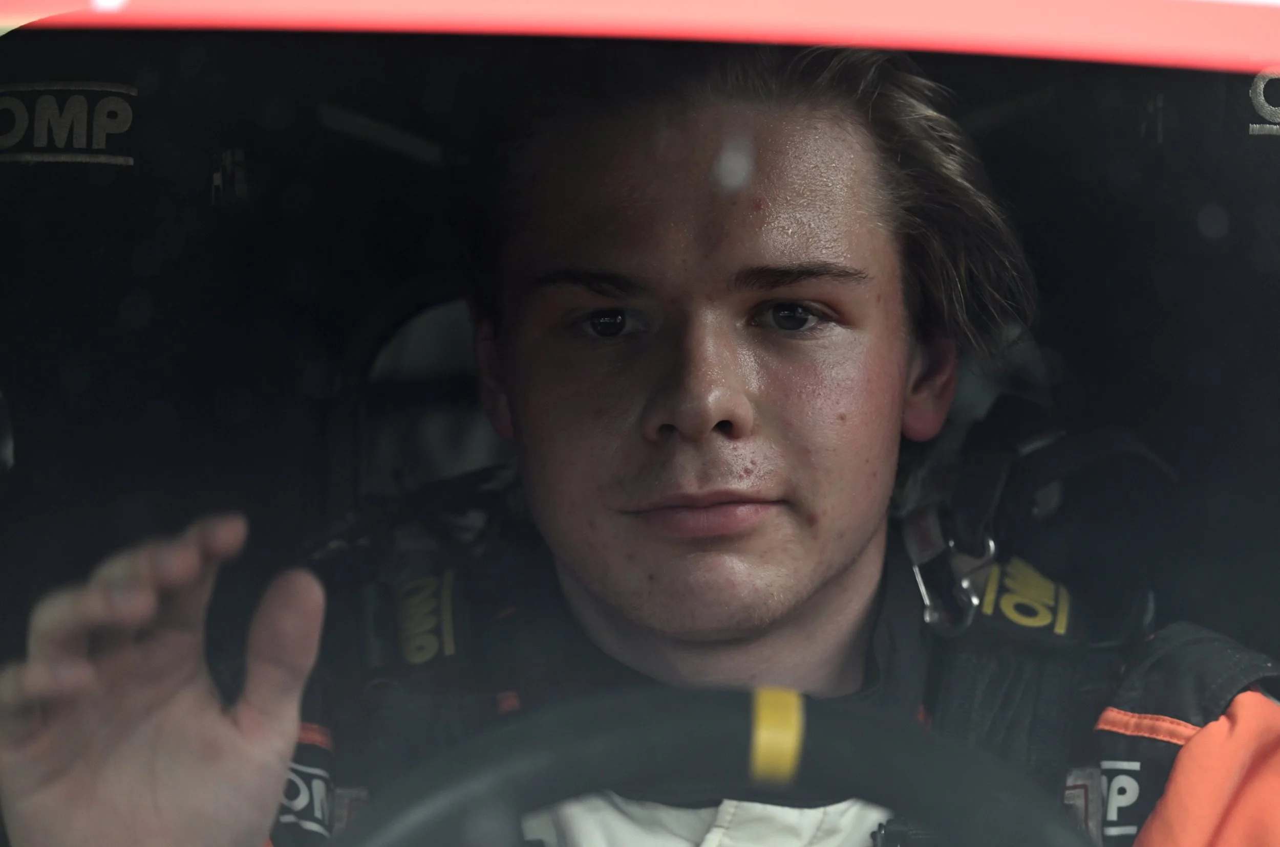 A young race car driver with wet hair and face, wearing a racing suit, looking through the windshield of a race car.
