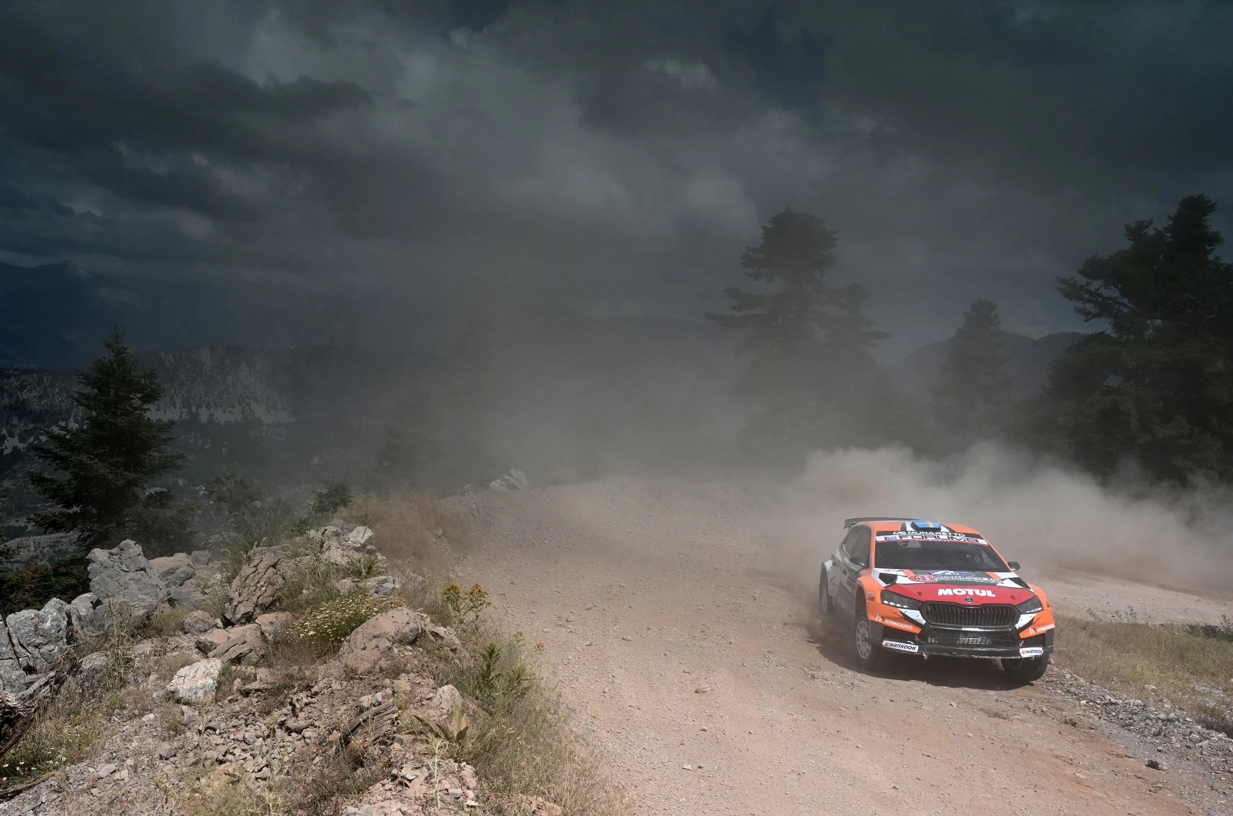 A rally car racing on a dirt road in a mountainous, forested area with dark, cloudy skies overhead.