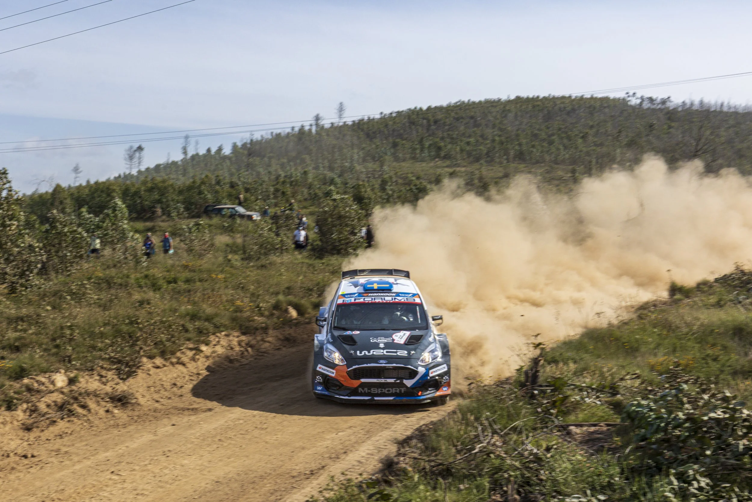 Race car driving on a dirt rally course, kicking up dust, with spectators and vehicles on the hillside in the background.