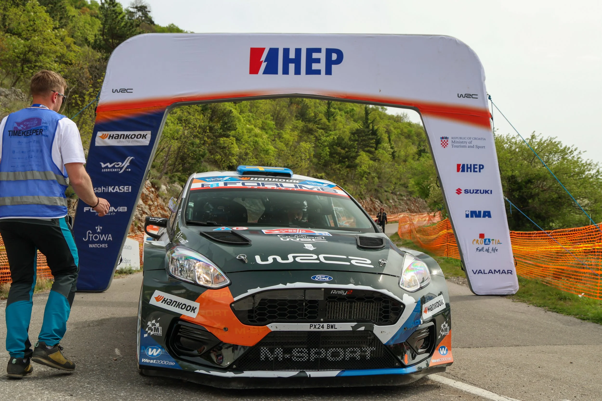 A rally car with sponsor decals, positioned under an archway with various sponsor logos, and a person in a blue vest standing nearby with trees in the background.