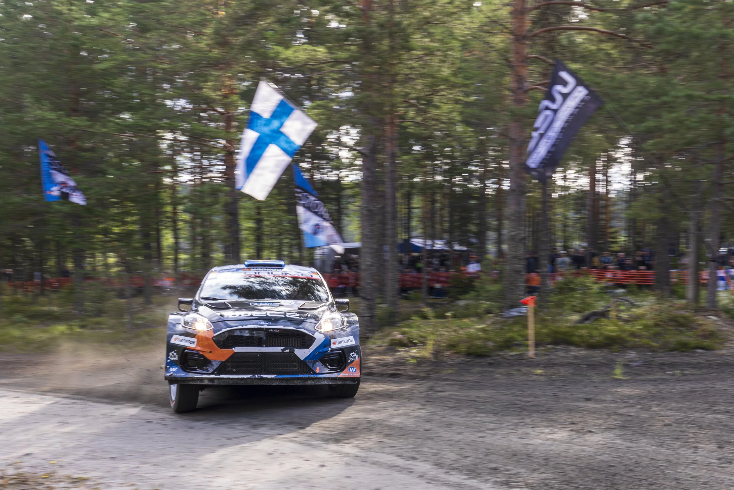 A rally car racing on a dirt track through a wooded area, kicking up dust behind it. There are flags flying in the background and spectators watching from the side.