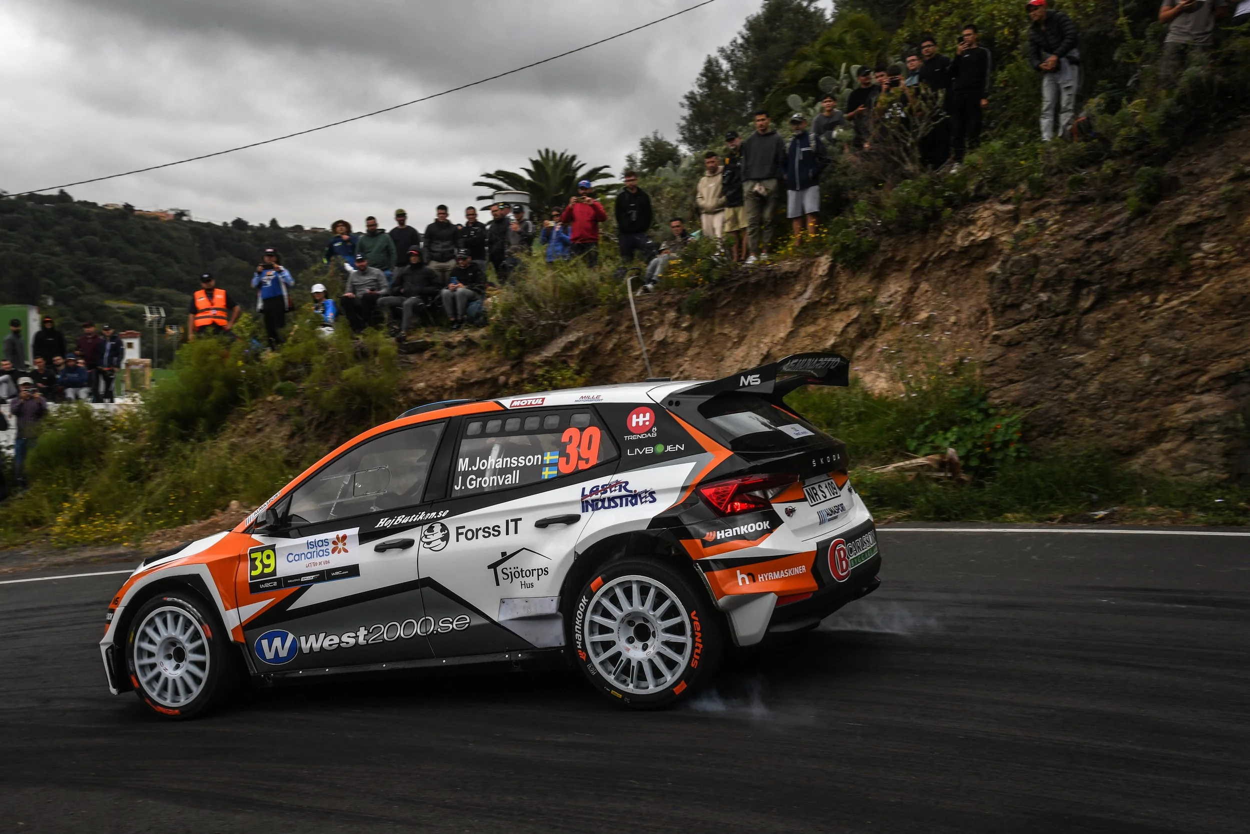 A rally car speeding on a wet mountain road with a crowd of spectators watching from the roadside and hillside.