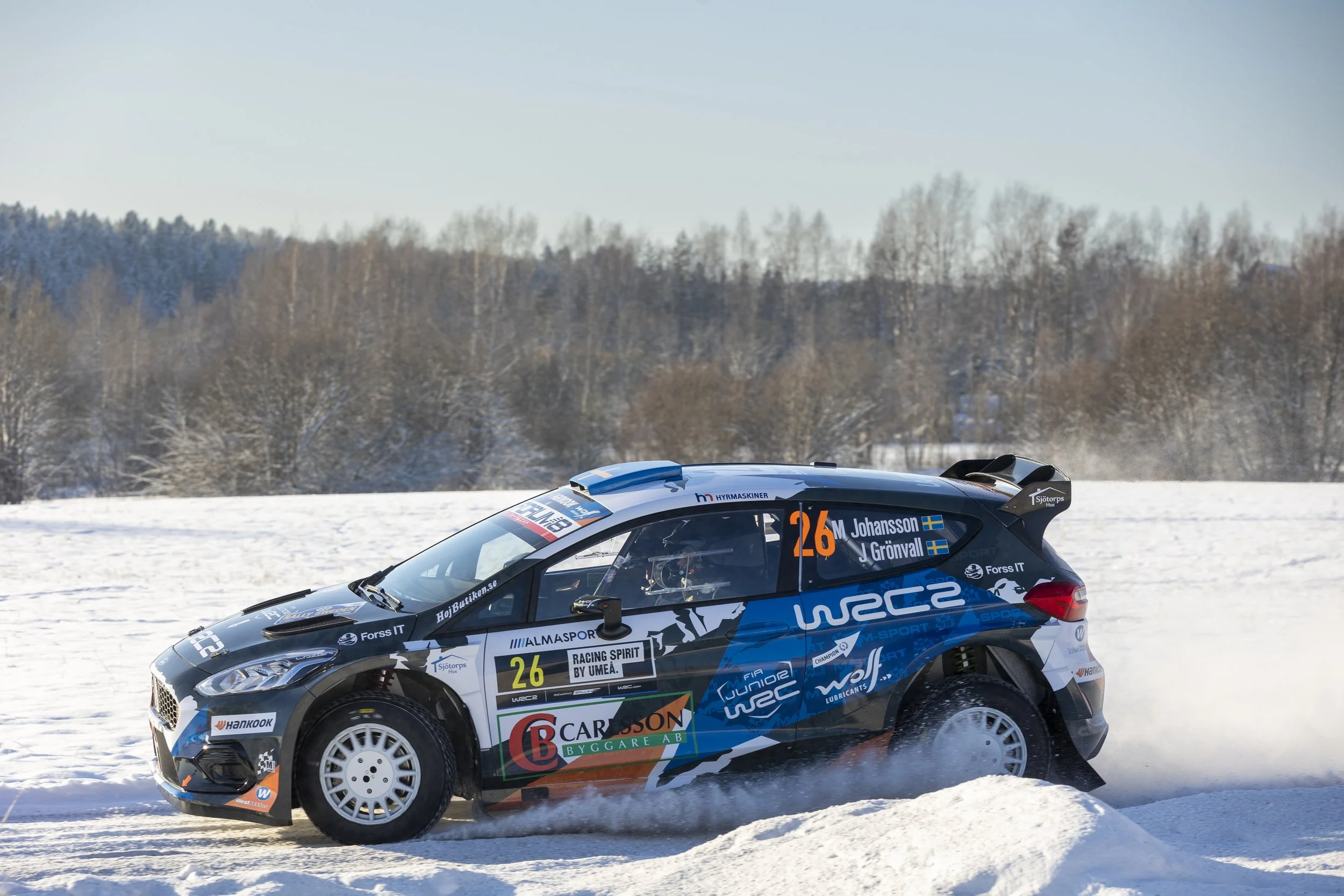 A rally car driving on snow-covered terrain with a background of snow-covered trees and a partly cloudy sky.