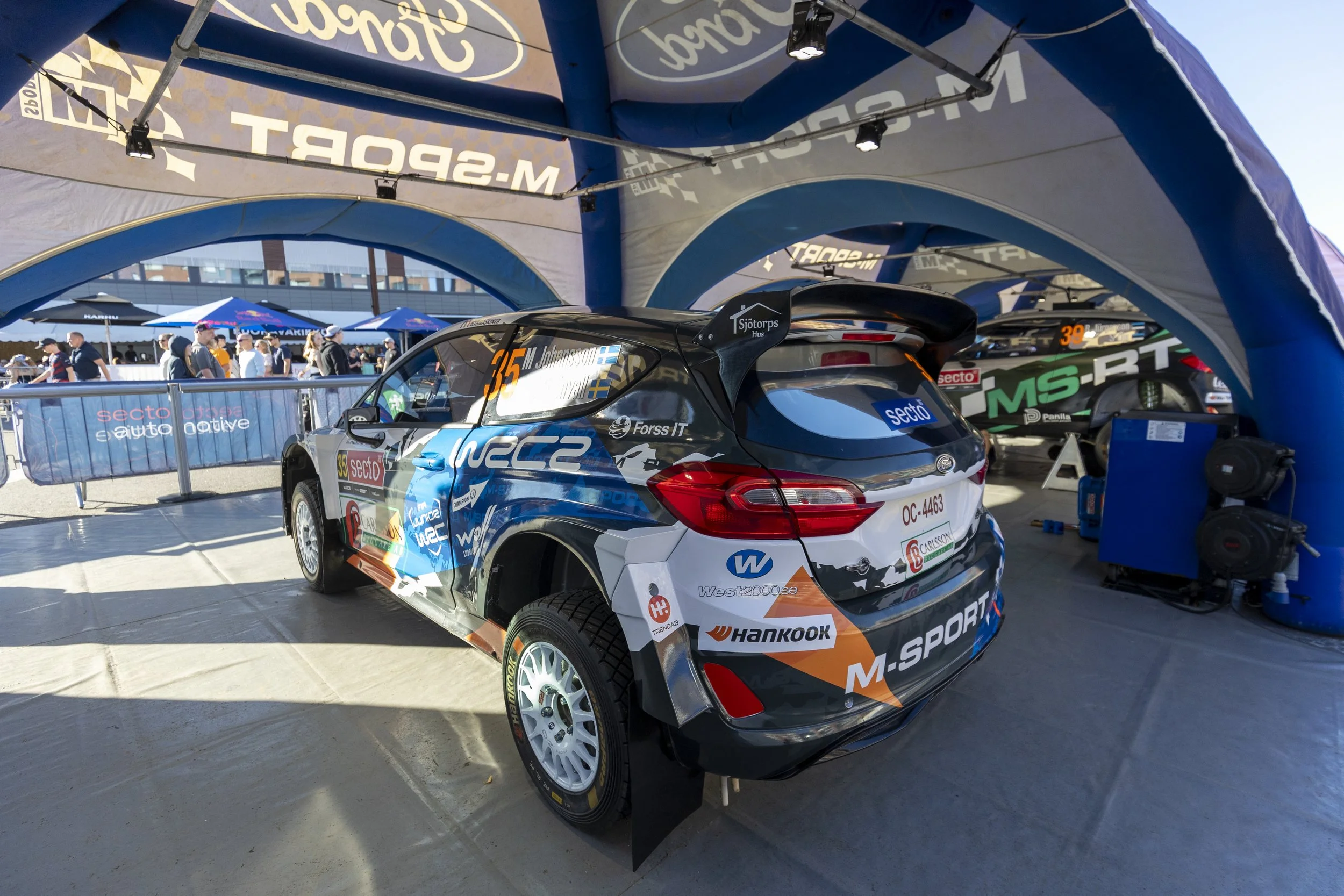 Race car with various sponsor logos parked under a blue tent at a motorsport event. People are visible in the background near the barriers and tents.