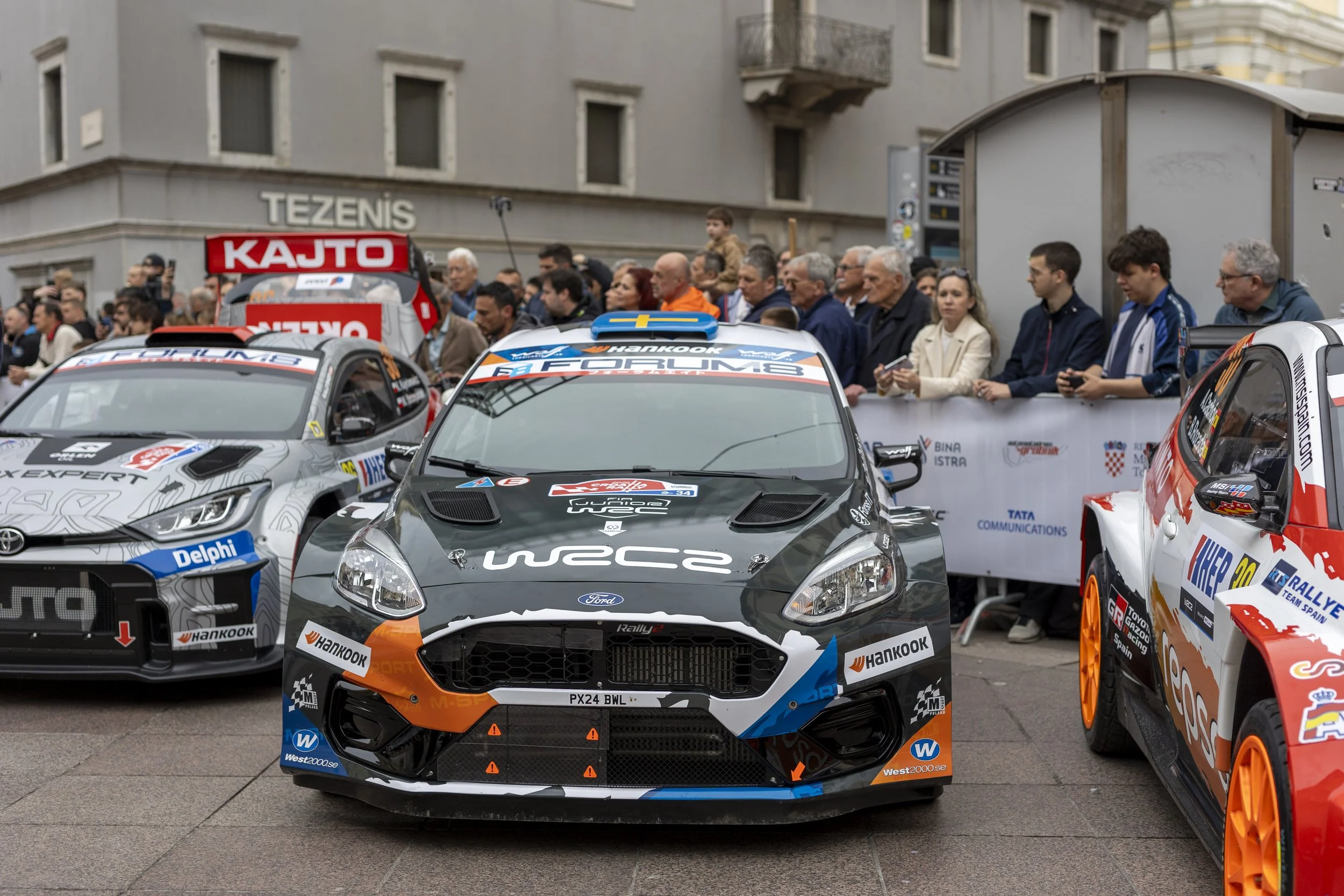 Race cars parked in a city square crowd of spectators lined up behind barriers watching, some taking photos, buildings in the background.