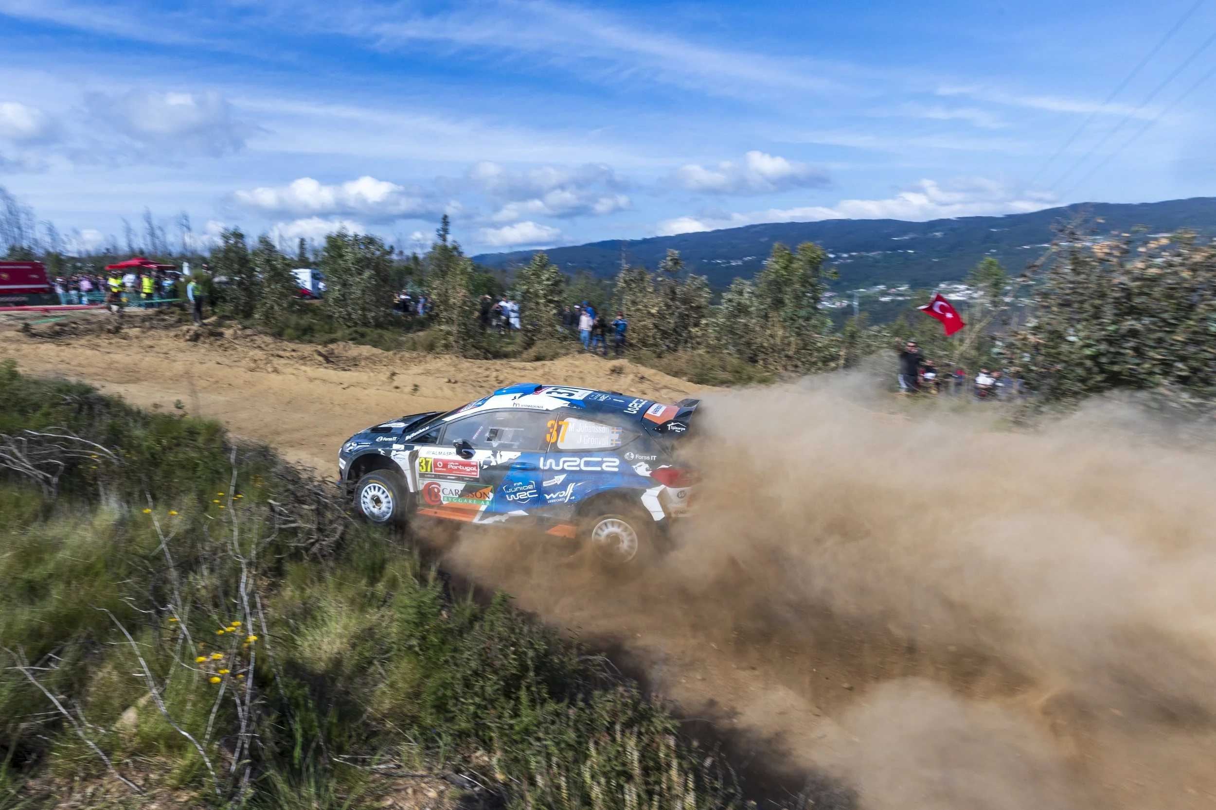 A rally car racing on a dirt track, kicking up dust, with spectators watching from the side, some holding flags, in a rural landscape with hills and a blue sky.