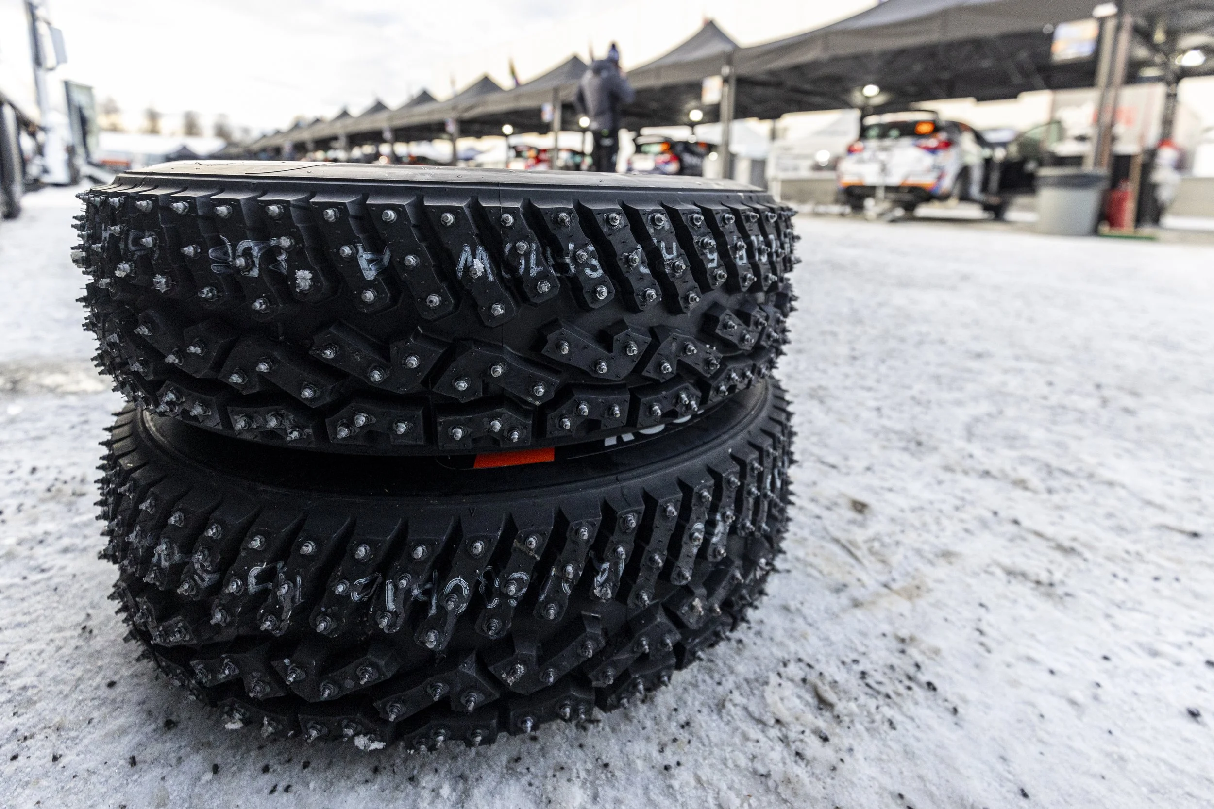 Stacks of black off-road tires with metal studs on a snowy surface, with a row of tents and parked cars in the background.