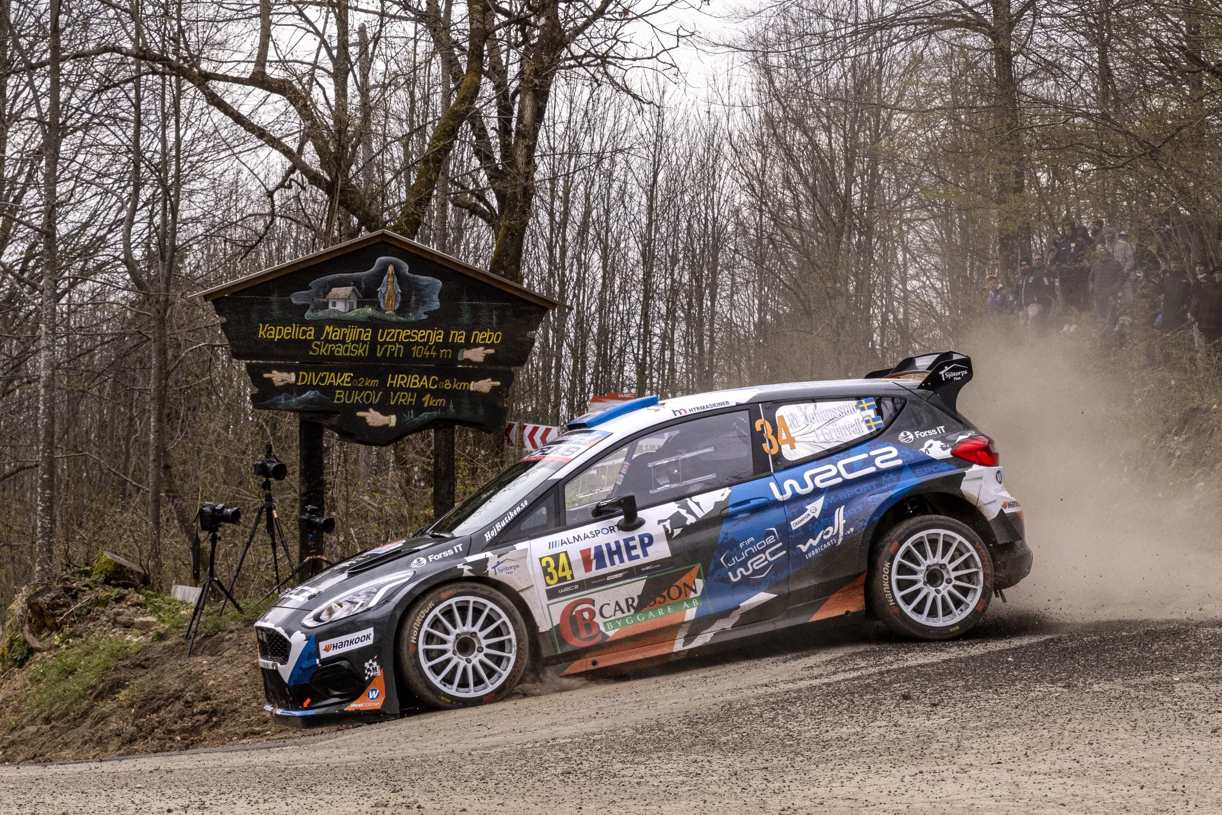 A rally car speeding on a dirt road in a wooded area, kicking up dust, with a group of spectators in the background, and a sign post with directions.