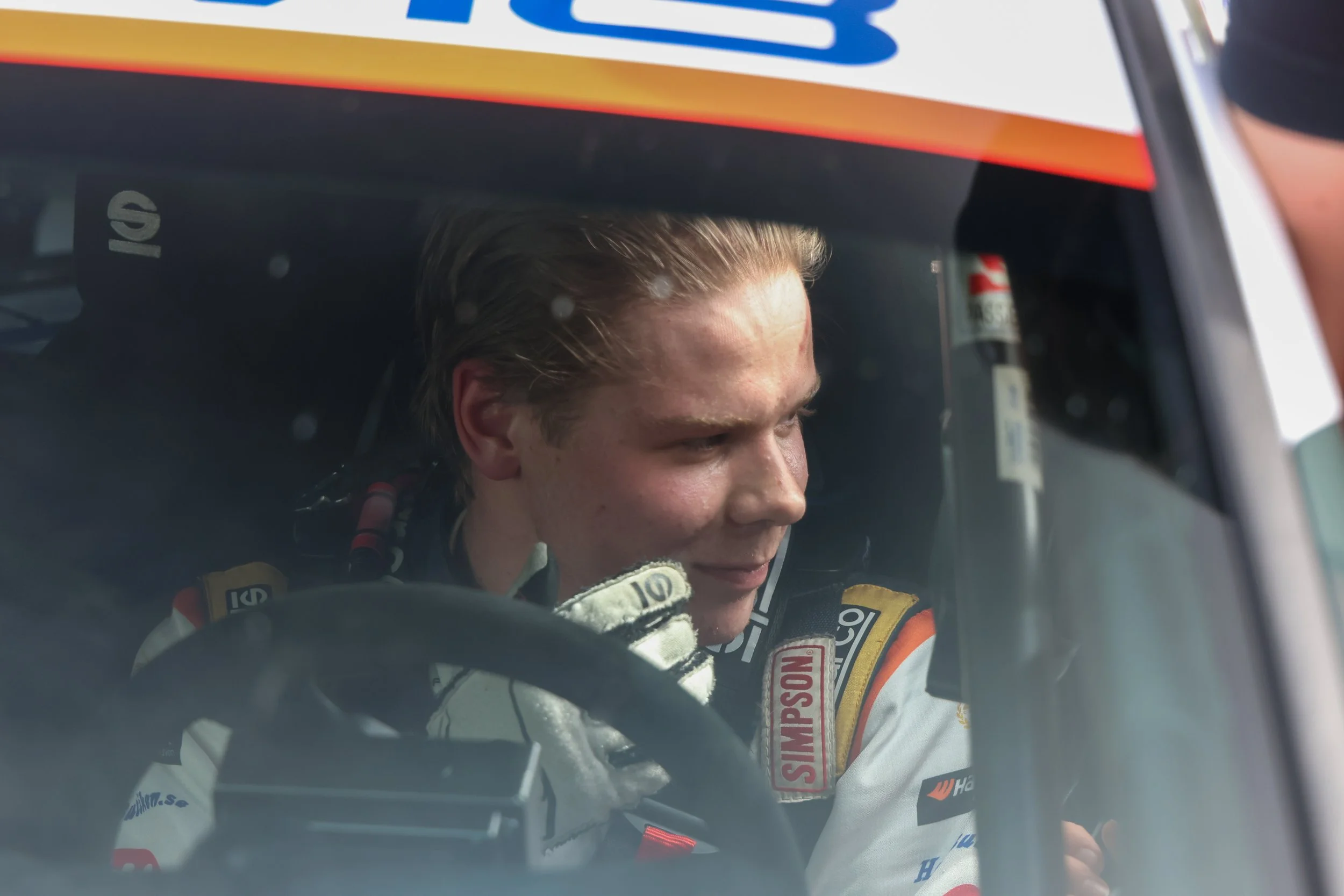 Close-up of a race car driver smiling inside the cockpit, wearing a white racing suit with sponsor patches, holding the steering wheel.