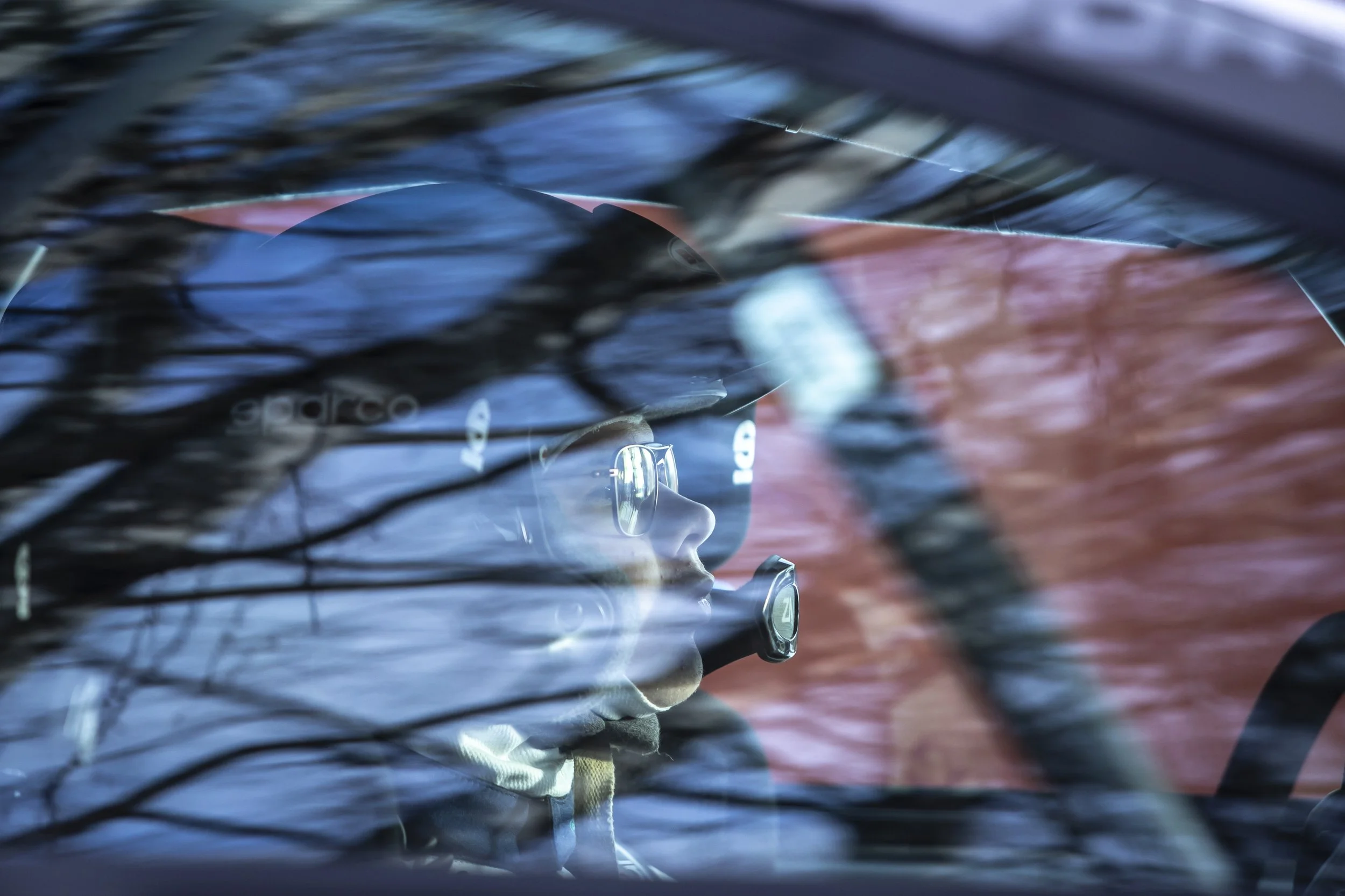 A person with curly hair and glasses driving a car, with reflections of trees on the windshield.
