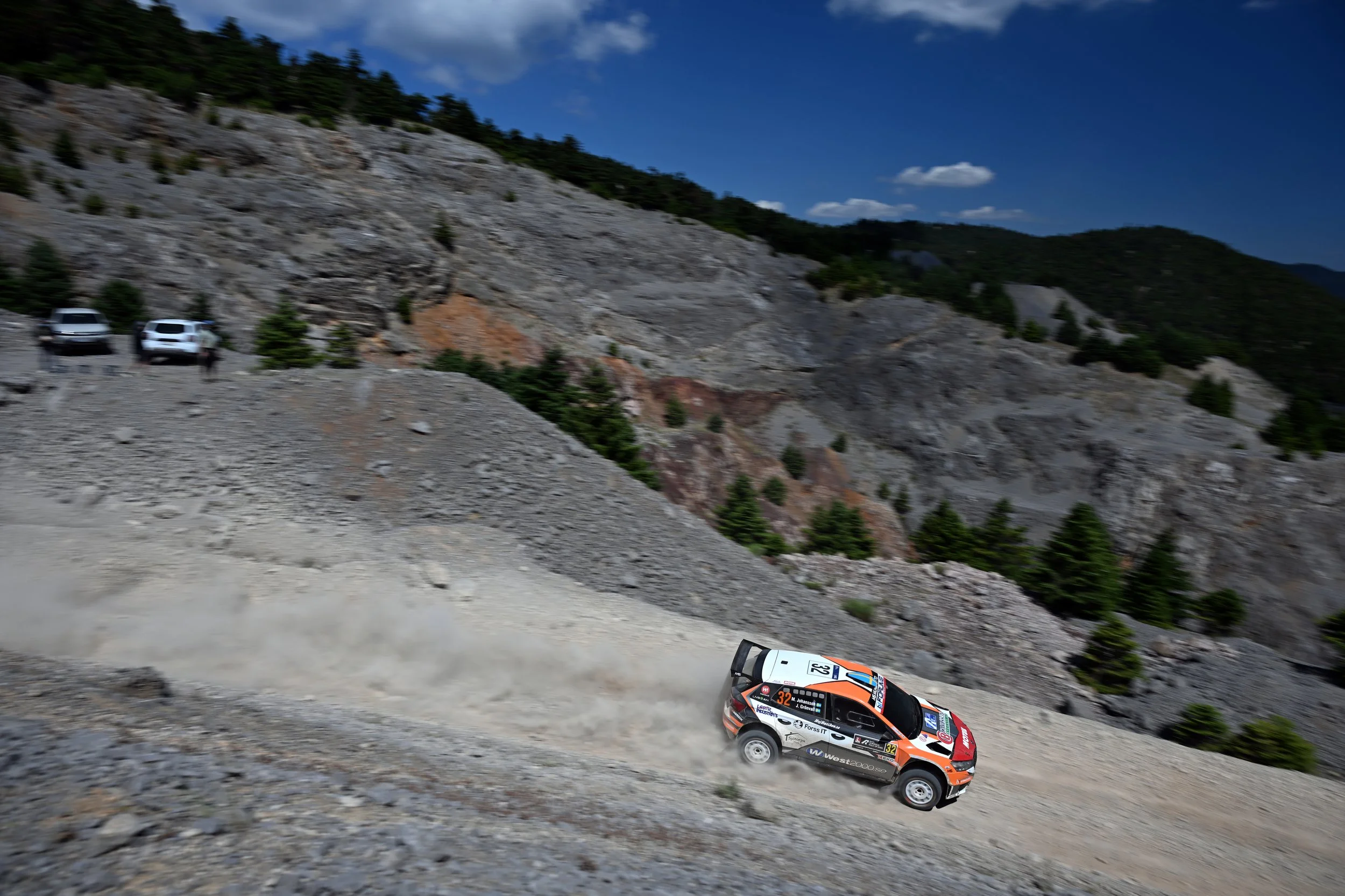 A rally car speeds along a dirt road in a mountainous area with rocky slopes and sparse pine trees under a blue sky with some clouds.