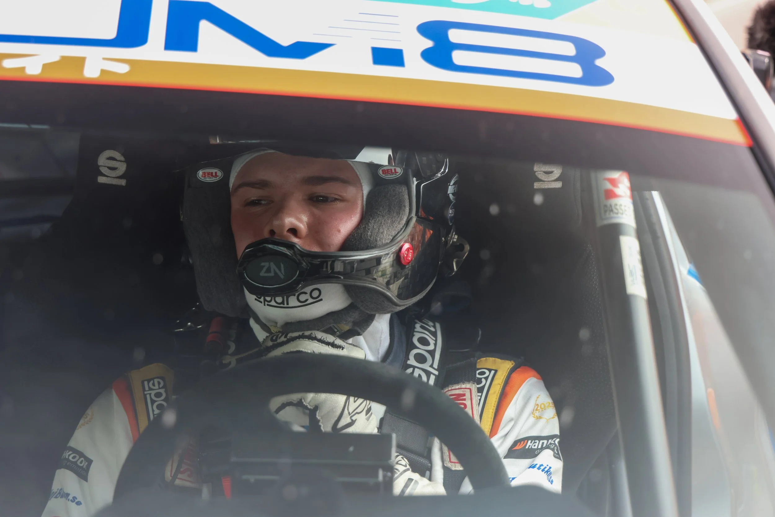 A race car driver wearing a helmet and racing suit, sitting in the cockpit of a race car, looking focused, with a blurred background.