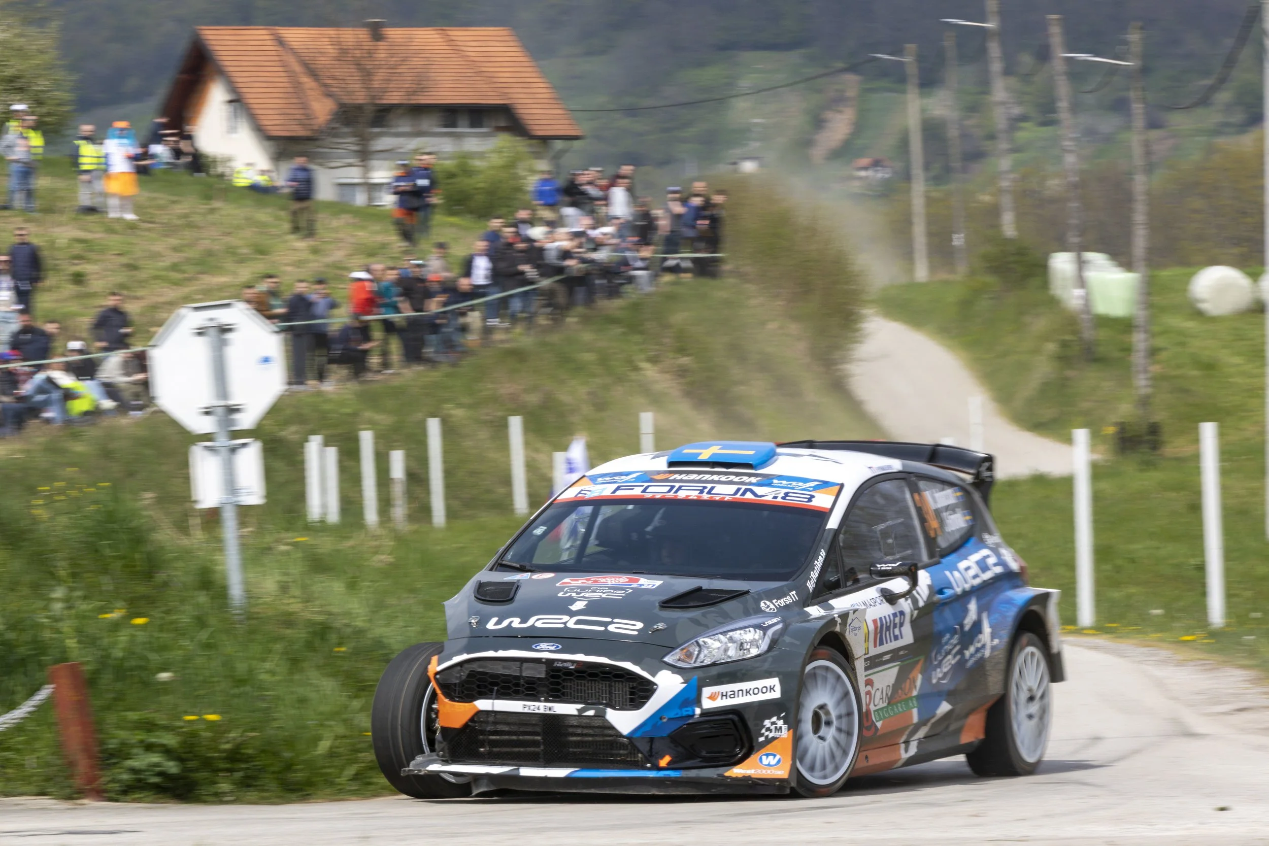 A rally car racing on a dirt road with a house and spectators watching in the background.
