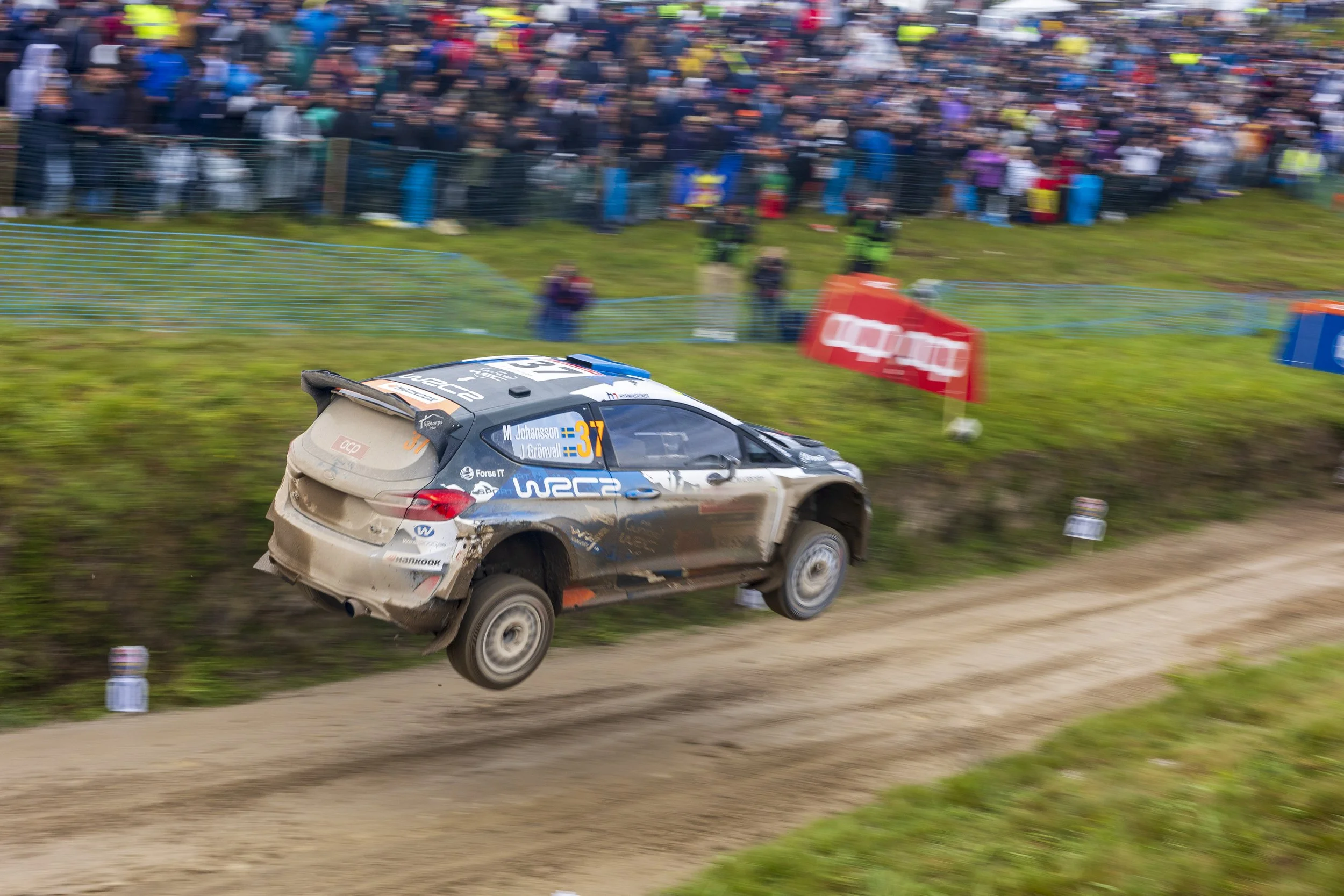 A rally car speeding along a dirt track with a large crowd of spectators behind a safety fence, some wearing colorful clothing, at a motorsport event.