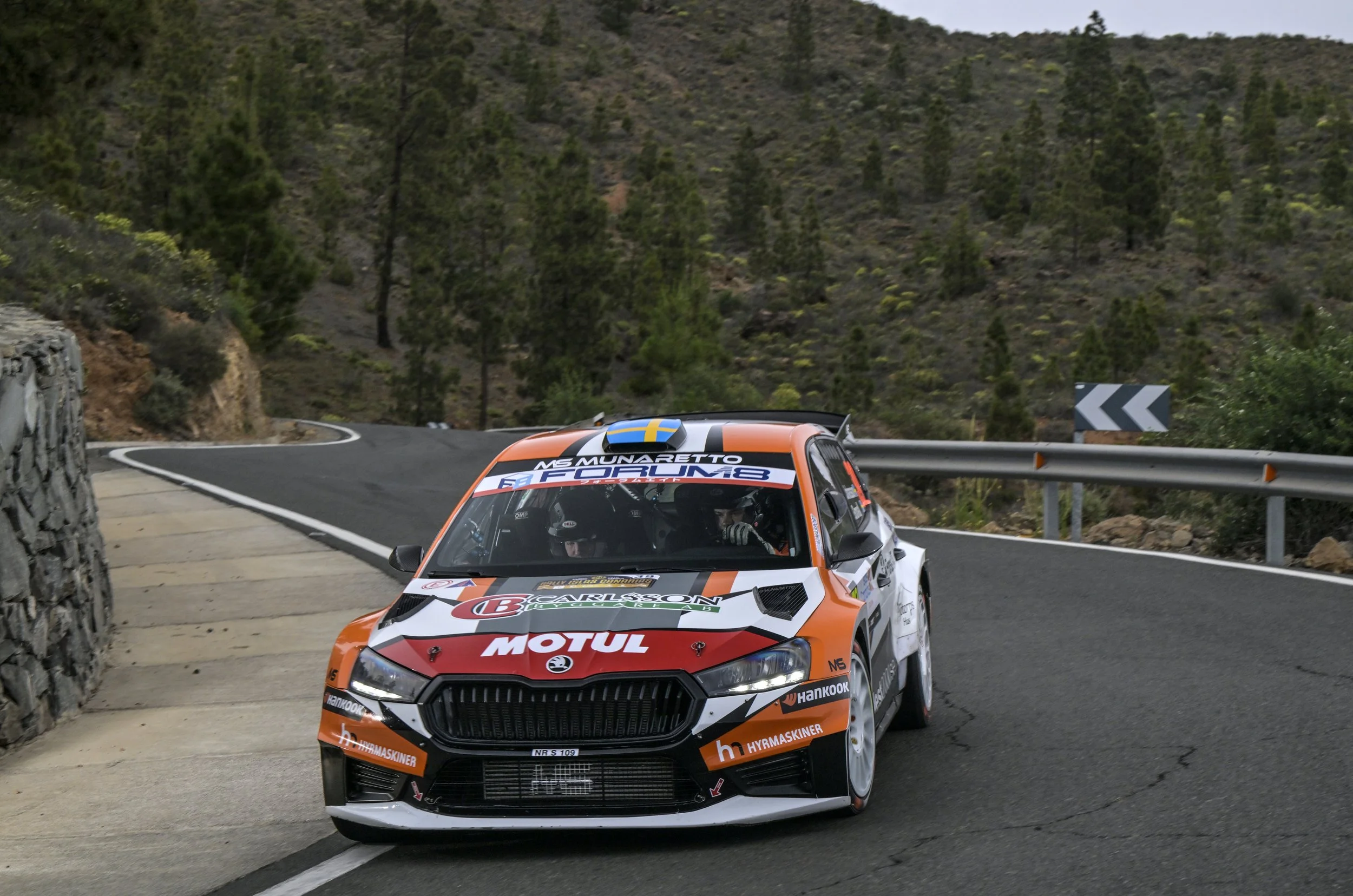 A rally car painted orange, white, and black navigating a mountain road with a guardrail, trees on a hillside, and a curve warning sign in the background.