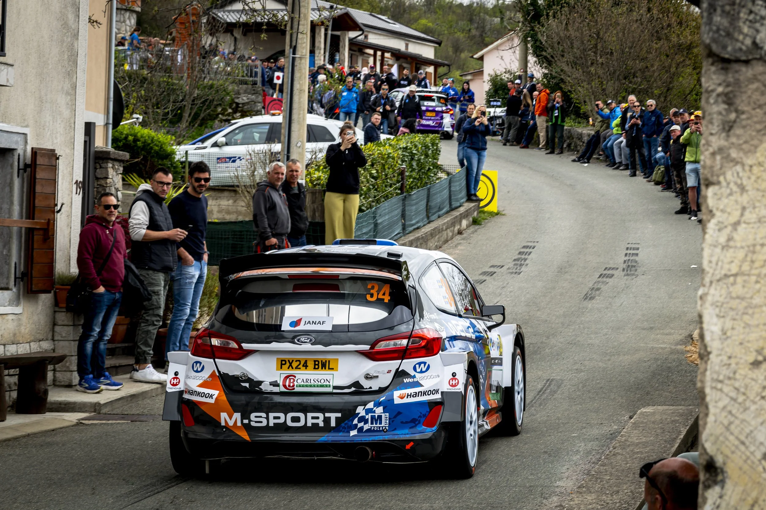 A rally car with sponsor decals and the number 34 on the windshield is racing along a narrow street lined with spectators. The crowd, some taking photos, watches as the car speeds past. The scene is set in a small town with houses and greenery.