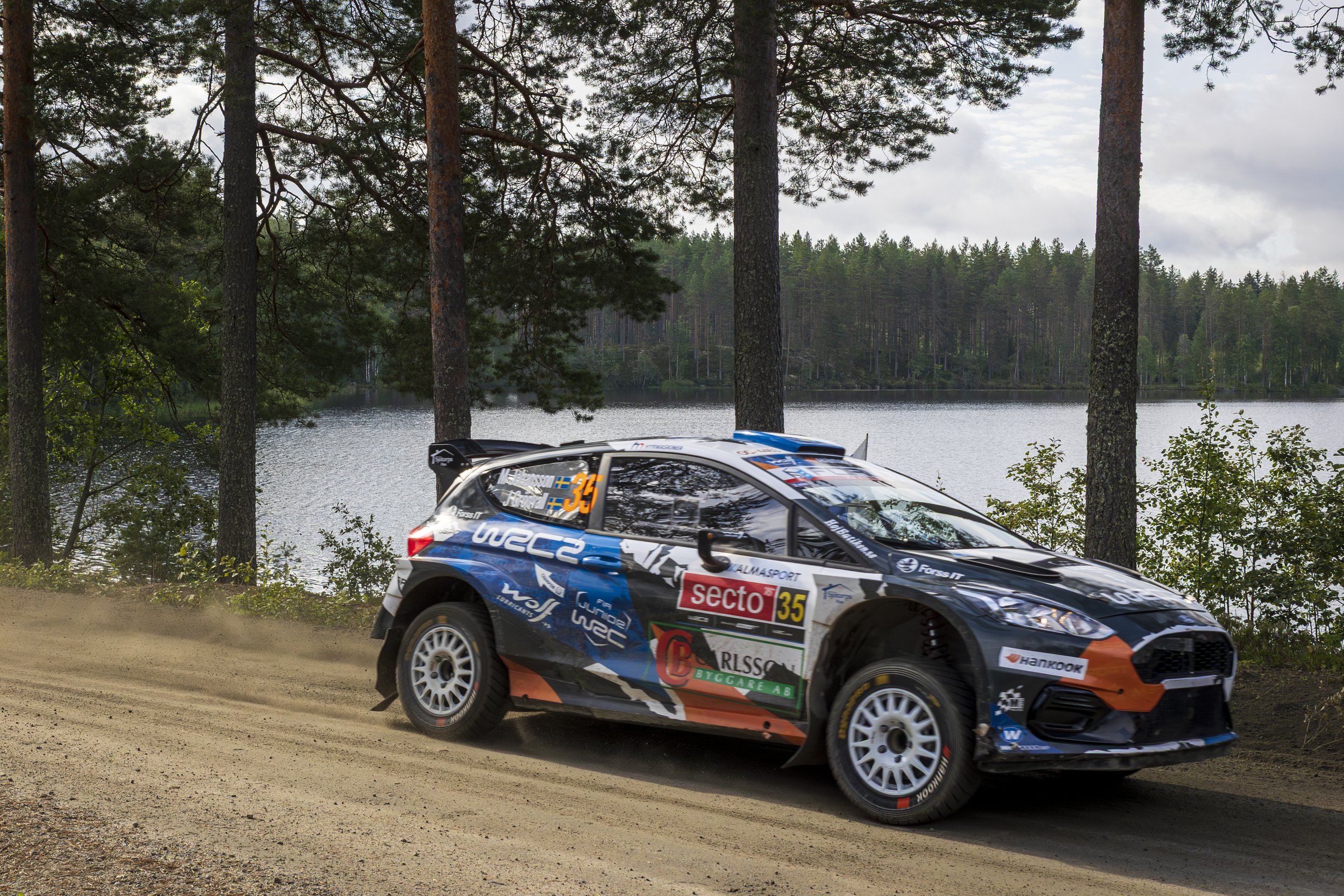 A rally car racing on a dirt road near a lake, surrounded by trees and overcast sky.