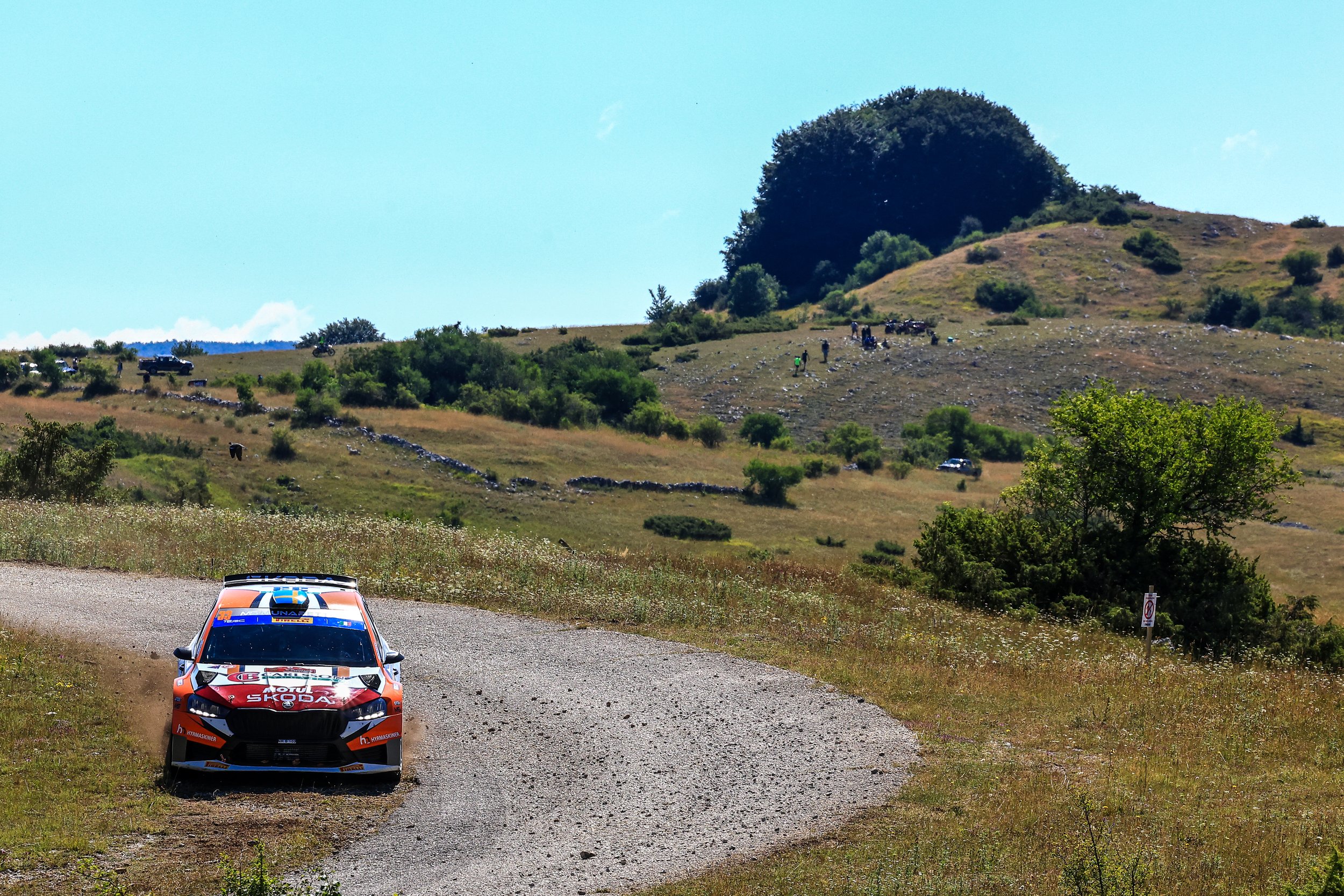 A red and white rally car on a gravel road in a hilly, rural landscape with green trees and shrubs and a large hill in the background.